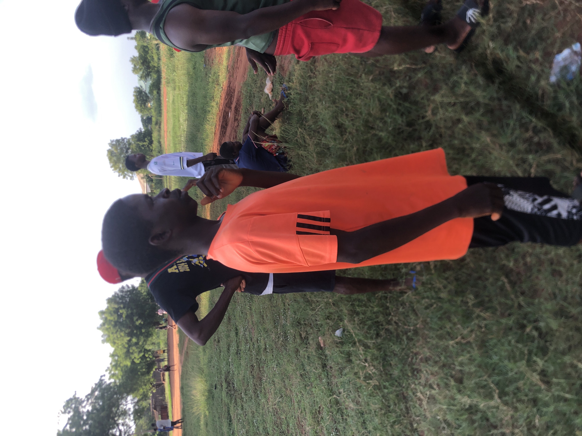 This media file captures an outdoor scene in Nsugbe, Nigeria, likely during the late afternoon or early evening under a bright, possibly overcast, sky. The setting is an open, undeveloped field characterized by patches of green grass interspersed with reddish-brown dirt paths.
In the immediate foreground, a young person is prominently featured in profile, wearing a bright orange short-sleeved t-shirt with distinct black stripes on the sleeves, indicative of a sports apparel brand. This individual is looking towards the left side of the frame, with their right hand raised near their mouth, possibly holding or gesturing with something.
Behind this individual, several other people are visible, suggesting a casual gathering or outdoor activity. One person stands prominently in a green sleeveless top and red shorts. Another is seen standing in a white t-shirt with a green logo (possibly a sports brand) and dark shorts. Further back and slightly to the left, a person is seated on the ground, wearing a blue top, with some light-colored string or material around their legs. In the background, numerous other figures are scattered across the field, some standing, indicating a larger group engaged in an unspecified communal or recreational activity. One person in a black t-shirt, seen from behind, has a colorful graphic that includes the partial text "202x." A black sandal or shoe with a distinctive white skeletal-like design is visible on the ground near the top right.
The overall atmosphere appears relaxed and informal. Trees line the distant horizon. A discarded plastic bottle is visible on the ground. No strong emotions are discernible, but the scene conveys a sense of community or shared leisure time in an outdoor environment.