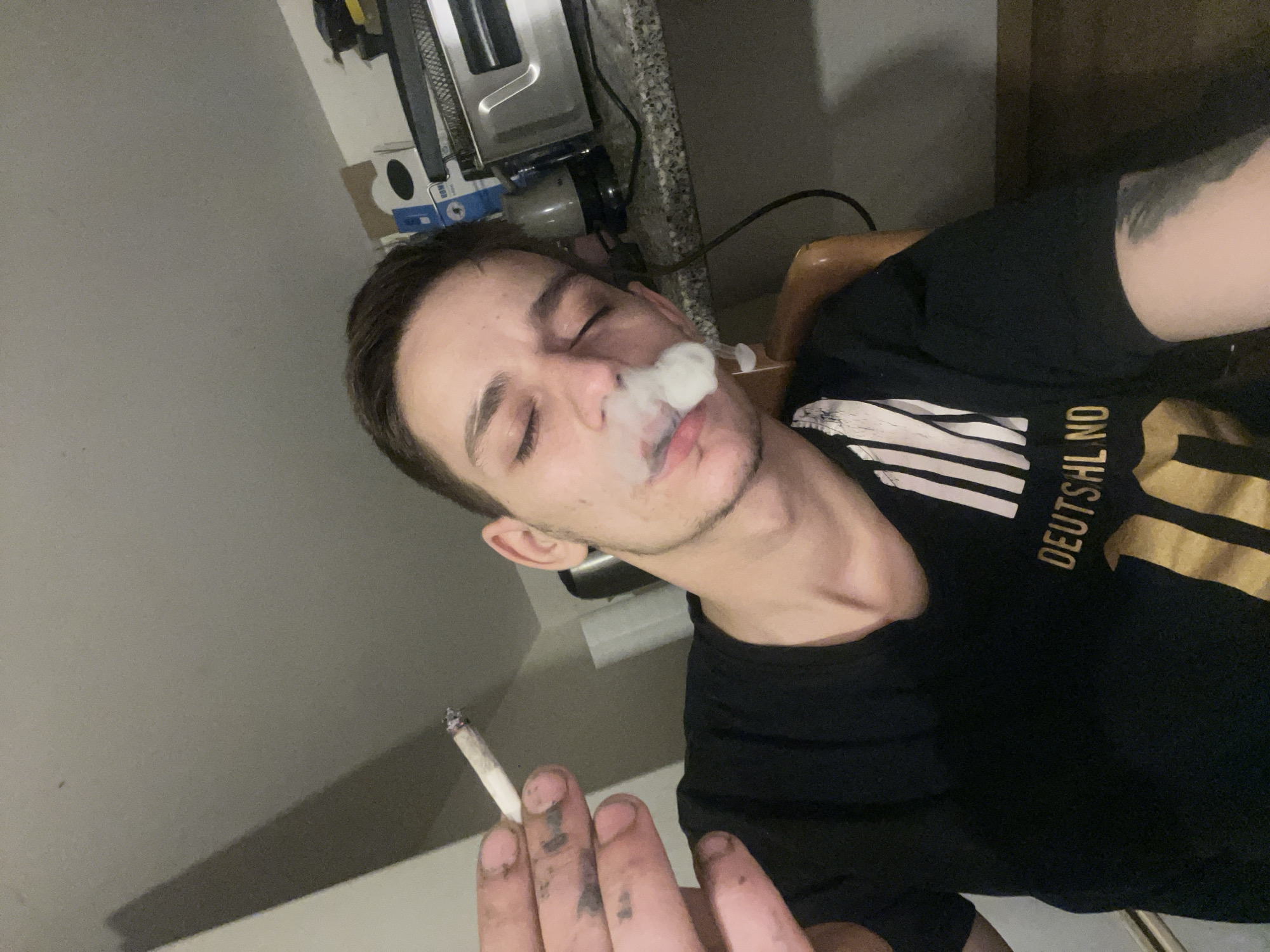 A young man is shown smoking a cigarette indoors. He is wearing a black t-shirt with a graphic design. The background includes a countertop with kitchen appliances and boxes. The lighting suggests an indoor environment, possibly a kitchen or living area. The overall impression is a candid snapshot of a moment.
