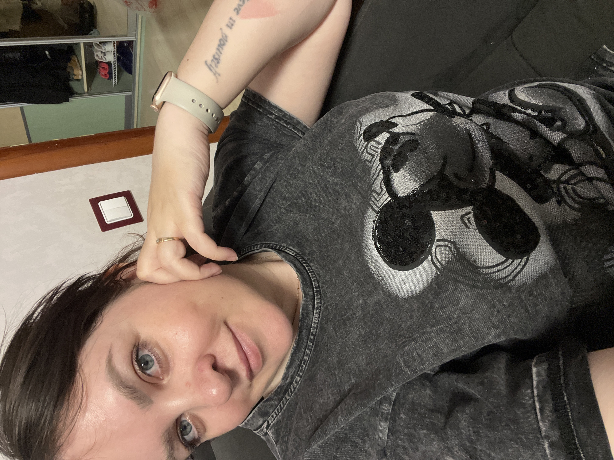 The image is an indoor selfie of a young adult woman, likely taken in a residential setting in Perm, Russia. She looks directly at the camera with a calm, slightly smiling expression. She has fair skin, dark hair, and light blue eyes.
The woman is wearing a dark gray, possibly acid-wash or faded, t-shirt. The shirt features a large, stylized design of a Mickey Mouse head, which appears to be embellished with black sequins or a shimmering texture. On her left wrist, she wears a light-colored (beige or tan) smartwatch with a rectangular face and a silicone band. A small, gold-colored ring with a stone is visible on her right ring finger, which is resting near her ear. A tattoo on her inner left forearm clearly reads "believe in yourself" in a cursive script.
The background shows parts of a room. Directly behind her is a dark-colored sofa or bed. To her left, a light-colored wall features a light switch with a white button encased in a dark red or brown frame. Further back, a metal shelving unit or clothes rack is visible, holding various items like clothes and shoes in shades of black, brown, red, and white. The wall behind the shelving unit is a light green or teal color, and a light brown surface, possibly a floor or another piece of furniture, is visible below. The overall impression is one of a casual, relaxed moment at home during the daytime.