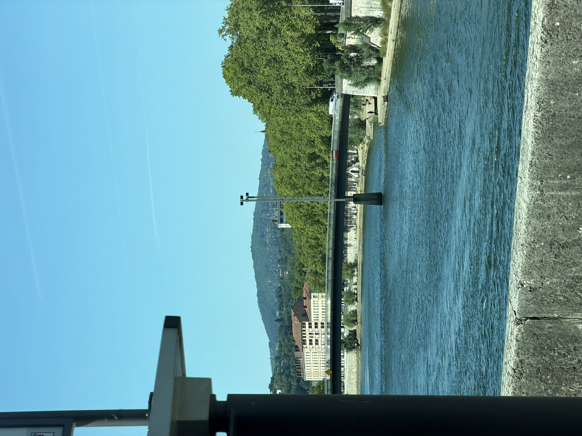 The image captures a view across the Saône River in Lyon, France, with a bridge spanning the water. In the foreground, a portion of a concrete barrier is visible, with a dark cylindrical structure on the left partially obscuring the scene. The Saône River is a vibrant blue, with ripples indicating gentle movement.
The bridge, a prominent feature, is a long, dark structure supported by concrete pillars. A white van is observed traveling on the bridge from left to right. On the far bank of the river, a multi-story building with a terracotta-tiled roof and many windows is situated, framed by lush green trees that dominate the midground. Beyond the trees, a verdant hill rises, dotted with buildings and more trees, suggesting the city's integration with its natural surroundings. The sky is a clear, bright blue, with faint white lines possibly indicating contrails from aircraft. The overall scene depicts a sunny day in an urban environment characterized by a river, a bridge, and architectural elements softened by abundant greenery.