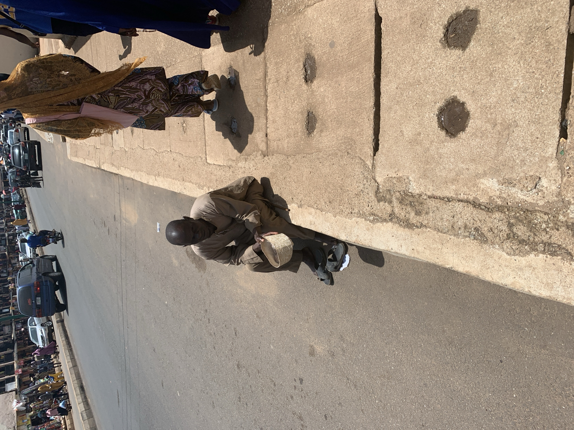 A man sits on the edge of a concrete sidewalk in Jos, Nigeria, holding a woven bowl in his lap. He wears a light brown, long-sleeved garment and dark sandals. His feet are bare, and he appears to be begging. Behind him, the sidewalk is made of large concrete slabs with some indentations and dark patches. A busy street runs alongside the sidewalk, with cars and motorcycles visible. Several people walk along the sidewalk and street, including a woman in a yellow headscarf and patterned dress, and another person in a blue garment. Buildings with storefronts line the far side of the street. The scene appears to be a bustling urban environment under bright sunlight.