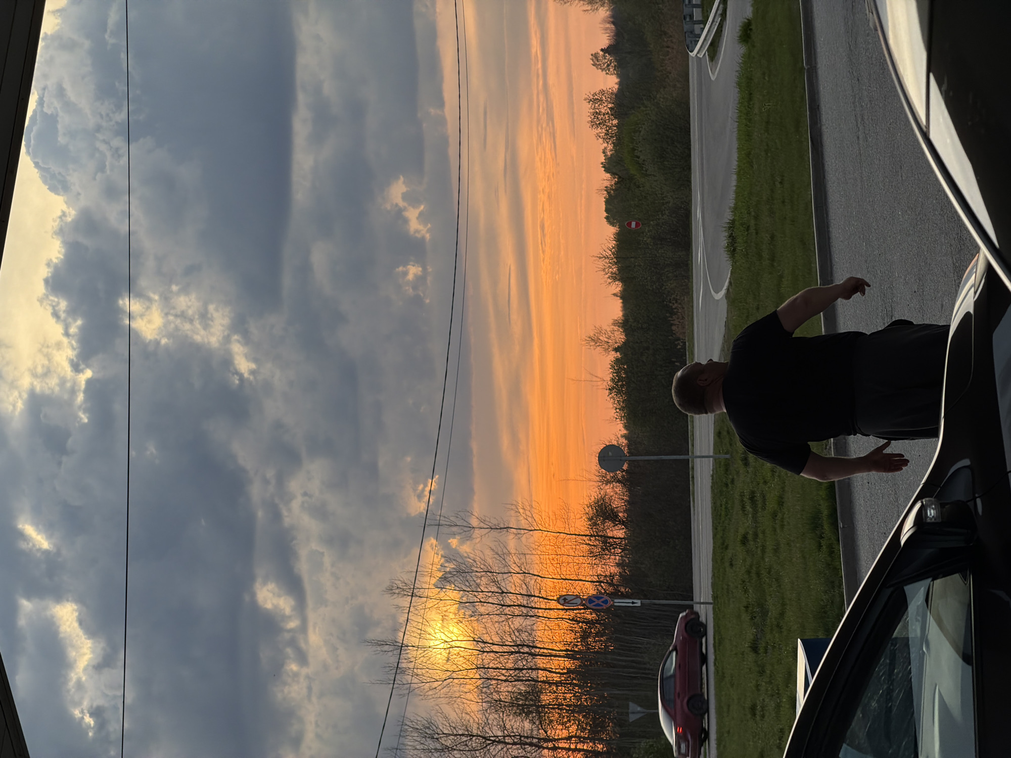 The image shows a man standing next to a car in Ostrov, Russia. He is facing a sunset with orange and gray clouds visible in the sky. There are trees in the background, and a road with a roundabout to the right of the man. A red car is parked on the side of the road near a road sign. Overhead power lines are visible cutting through the sky. The man is wearing a dark shirt and pants, and he has short hair. He is standing near the driver's side of the car, and one of his hands appears to be outstretched. The environment appears to be at dusk or dawn, given the lighting conditions.