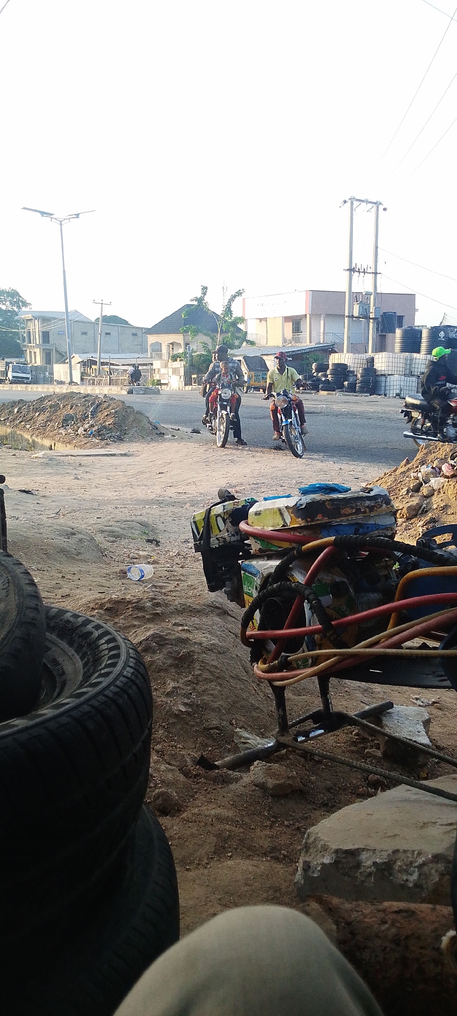 The image depicts an outdoor scene in Lokoja, Nigeria, at an apparent intersection. Two men ride motorcycles toward the camera, while another motorcyclist with a green helmet sits on their bike further back. Behind them are buildings, including a building with black tanks stacked against its wall, and utility poles. Piles of dirt and rubble are scattered around the intersection. In the foreground, stacks of tires and a piece of machinery with multi-colored hoses dominate the view, partially obscuring the broader scene. An empty plastic bottle lies on the ground near the tires. The sky is overcast, suggesting diffused light.