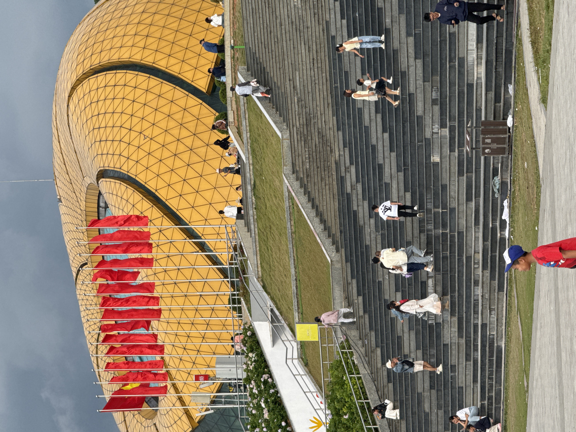 The image depicts the exterior of the Da Lat Cultural Palace in Da Lat, Vietnam. The building features a distinctive, undulating golden facade with a patterned geometric design. A long, wide staircase leads up to the building, with numerous people walking up and down the steps.
In the foreground, a young boy wearing a red shirt and a blue and white baseball cap is looking to the right. The staircase itself is constructed with dark gray stone steps and lighter gray risers, with metal railings on either side.
Several individuals and groups are visible on the stairs. On the lower steps, a woman in a white dress and a young child in dark shorts are walking up. To their right, a young girl in a light yellow top and blue jeans is descending. Further up the stairs, a man in a white t-shirt and black pants is walking down. On the middle section of the stairs, a woman in a white dress and a person in a light blue top and dark pants are also ascending. A man in a gray shirt and black pants is walking up a higher set of stairs.
On the upper levels, closer to the building, several people are seated or standing. A group of people are gathered on a grassy area to the left of the stairs, near some flowering bushes. A line of red flags, some with yellow stars, are prominently displayed on flagpoles in front of the golden building. The sky above is overcast and appears gray. The overall scene suggests a public gathering or a day of activity at the cultural center.