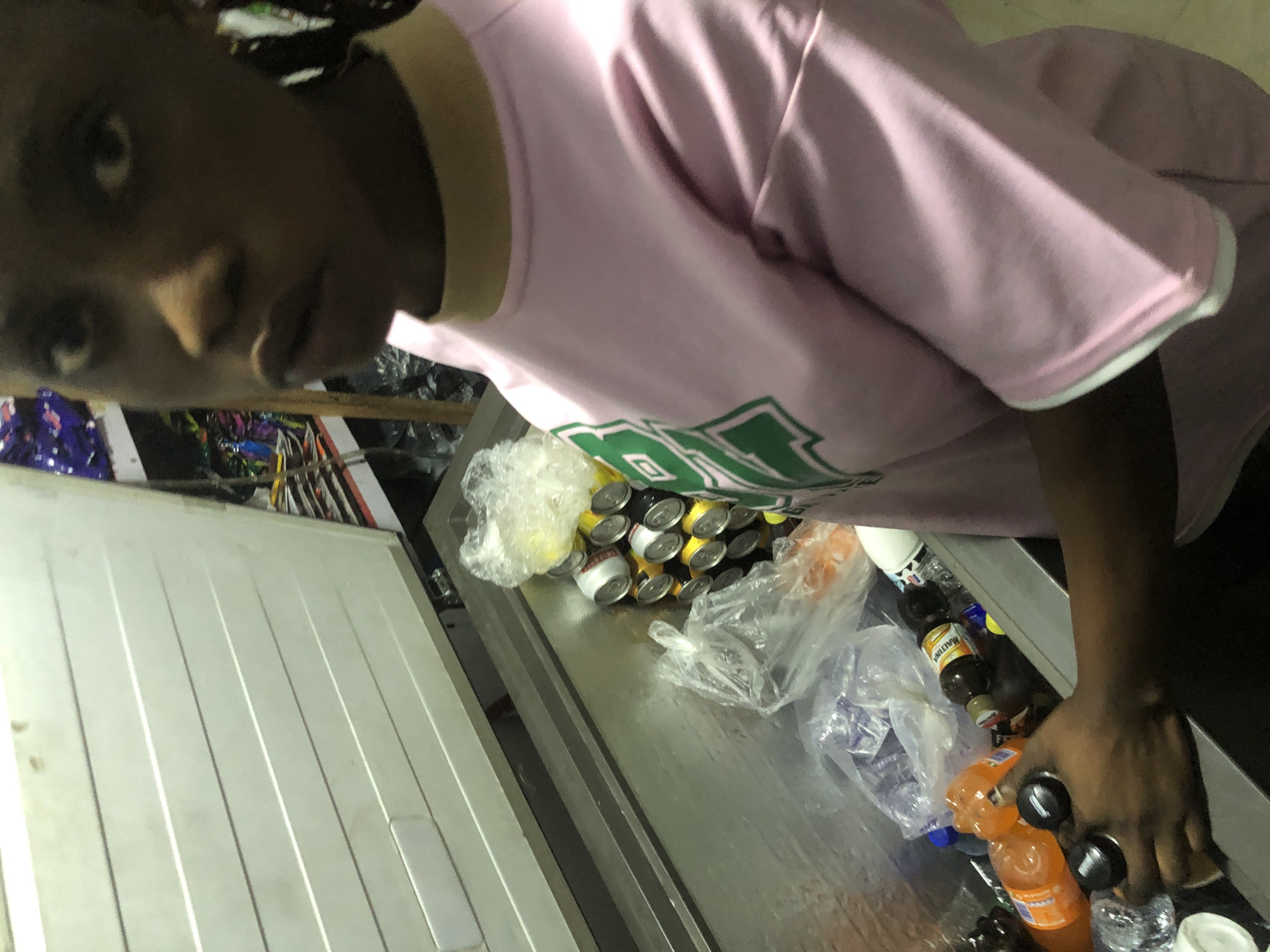 In Ado, Nigeria, a young woman wearing a pink t-shirt with green lettering is standing next to an open, stainless steel chest freezer. The freezer contains several bottles of beverages, including orange soda and Maltina, as well as a pile of canned drinks, some wrapped in plastic. The woman's hand is reaching into the freezer, holding two dark-colored caps. The scene appears to be indoors, possibly a shop or a food storage area, with shelves of packaged goods visible in the background.