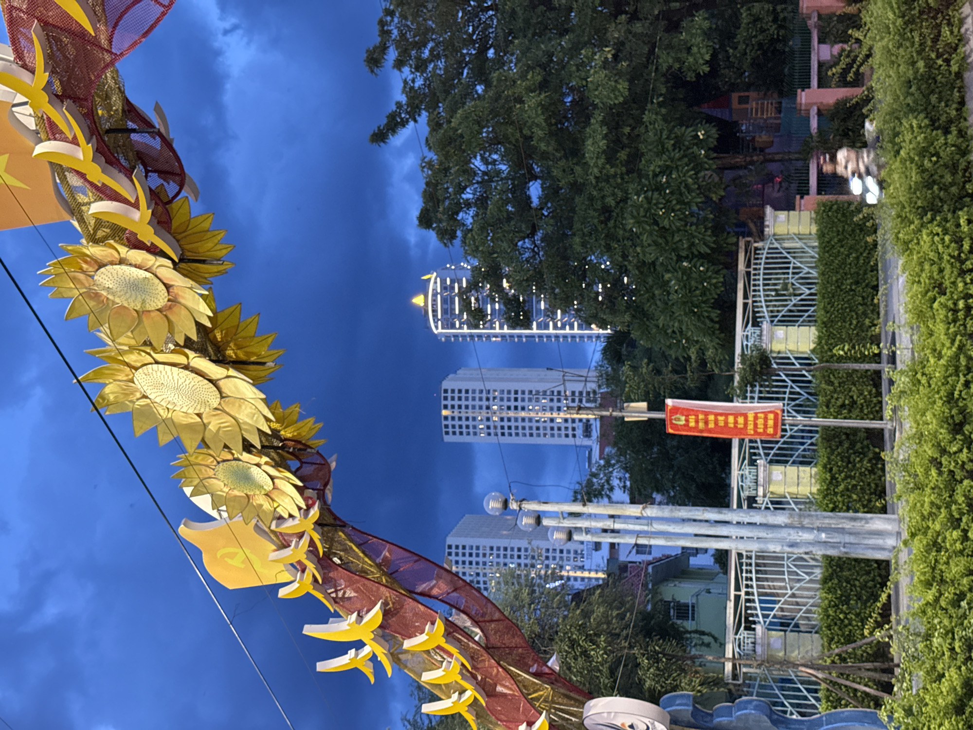 This is an image captured at dusk in Nha Trang, Vietnam. In the foreground, a decorative dragon-like structure adorned with large, golden sunflower-shaped ornaments and red and yellow streamers arches across the sky. Behind the structure, two tall, modern apartment buildings with illuminated windows stand prominently. Trees and a white fence line the street, with a red banner displaying Vietnamese text hanging from a pole. The sky is a deep, cloudy blue, indicating the transition from day to night. The scene suggests a festive or celebratory atmosphere, likely during a holiday or special event in the city.