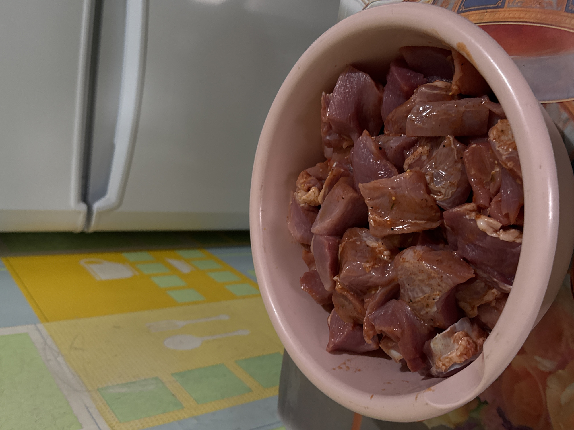 This indoor photograph, taken in Бородулино, Russia, provides a close-up view of raw, cubed meat in a light pink plastic bowl. The meat, cut into irregular pieces, appears to be well-seasoned with various spices, indicating preparation for cooking such as marinating or stewing. The bowl is angled slightly towards the camera, dominating the right side of the frame.
In the background on the left, a portion of a white kitchen appliance, likely a refrigerator or freezer, is visible, showing its vertical seam and smooth surface. Below this appliance, and extending into the foreground, is a patterned kitchen mat or floor covering. This mat features a colorful design in shades of yellow, green, and light blue, with white outlines of common kitchen utensils like a fork and spoon, along with other square shapes. To the right, behind the bowl, a small section of a vibrant, patterned surface, possibly a tablecloth or another decorative item, can be seen, adding a splash of warmer tones.
The scene depicts a domestic kitchen environment, characterized by the presence of cooking ingredients and kitchen utilities. The lighting suggests an indoor setting, likely with artificial illumination. No people are visible, and there is no discernible text within the image. The overall impression is one of active food preparation in a home setting.