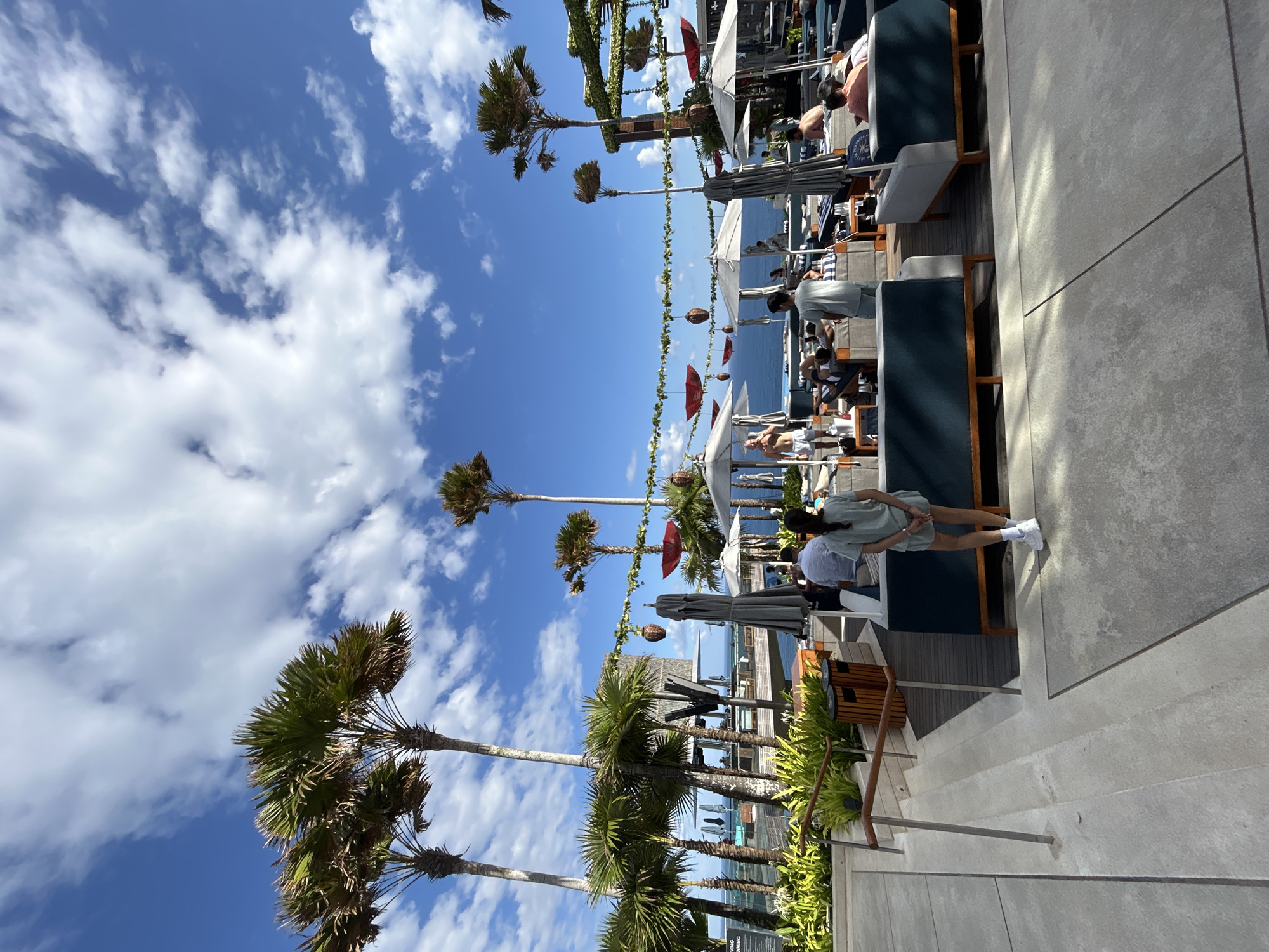 The image depicts an outdoor lounge area with a view of the ocean under a bright blue sky with scattered white clouds. Tall palm trees are a prominent feature, some swaying gently in the breeze. In the foreground, there are lounge chairs and sofas arranged around low tables, some occupied by people. A young woman in a light-colored casual outfit stands with her back to the camera, facing the lounge area. Umbrellas provide shade over some of the seating. In the background, more seating areas are visible, along with glimpses of the ocean and structures that suggest a resort or beach club. Decorative elements like garlands and hanging lanterns are strung across the area. The overall ambiance is relaxed and tropical. This scene is consistent with the popular beach clubs and resorts found in Kuta Selatan, Indonesia, known for their ocean views and vibrant atmosphere.