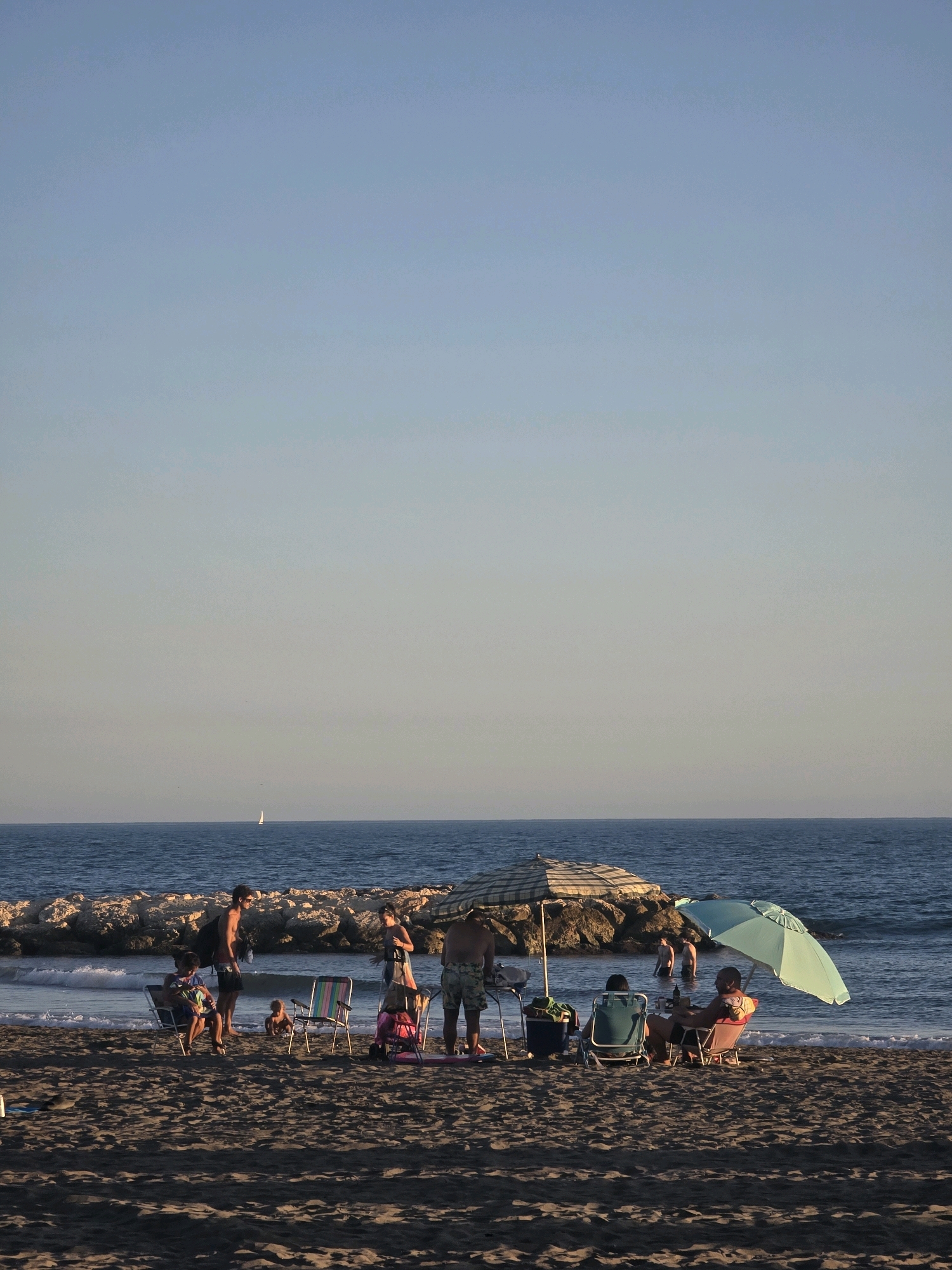The image depicts a beach scene in Málaga, Spain, with several groups of people enjoying a sunny day by the Mediterranean Sea. In the foreground, the sandy beach is populated with beach chairs, umbrellas, and a few individuals. One group, partially shaded by a checkered umbrella, includes two people seated in chairs and another standing. To the right, a light blue umbrella shades a person sitting in a chair. Further back, a cluster of people are gathered near the water's edge, with some standing and others seated. Children are visible playing in the sand. The sea is calm with gentle waves reaching the shore. A rocky jetty extends into the water on the left side of the frame, and a small sailboat is visible in the distance on the horizon. The sky is clear and pale blue.