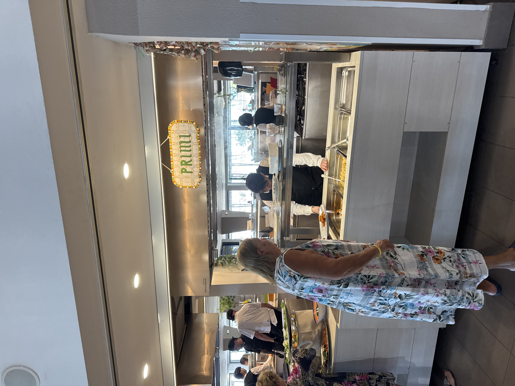 This is an image of a buffet line in what appears to be a restaurant or hotel in Isola delle Femmine, Italy. A sign above the buffet reads "PRIMI". There are several people visible, including guests and staff. On the left, a woman in a floral dress is looking at the food. Behind her, staff members in black uniforms are serving. In the center, a person in a black vest is serving food to a guest holding a plate. The buffet includes various food items in metal trays, such as pasta and what looks like meats or vegetables. The environment is indoors with bright lighting and a modern aesthetic.