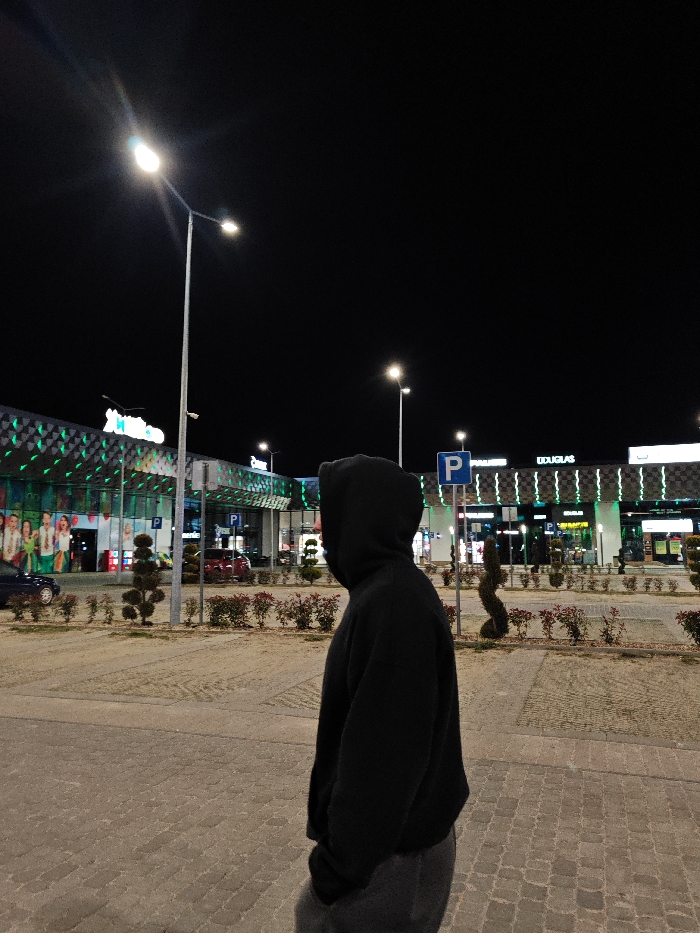 This nighttime image depicts a person in profile standing on a paved surface outside a retail park in Shumen, Bulgaria.
The central figure is a person, visible from the mid-torso, facing right. They are wearing a black hooded sweatshirt with the hood up, completely obscuring their head and face. Dark grey trousers are visible below the sweatshirt. The person stands on light-colored interlocking pavers, likely a pedestrian walkway or parking area.
In the background, two large commercial buildings are visible. To the left, a building features a prominent illuminated white "Metro" sign against a light blue background. Its facade is adorned with a dynamic green, grid-like LED lighting pattern. Below the "Metro" sign, figures (possibly mannequins or people) are visible through large windows. A dark blue car is partially visible parked in front of this building. To the right, another building displays an illuminated "DOUGLAS" sign and also features a similar green LED lighting pattern on its facade.
Between the person and the buildings, rows of small, manicured bushes with reddish foliage and several topiary-style shrubs are planted in landscape beds. Multiple tall, grey streetlights with bright, white LED lamps illuminate the scene. A blue parking sign ("P") on a pole is visible to the right of the central figure. The sky above is dark and clear.