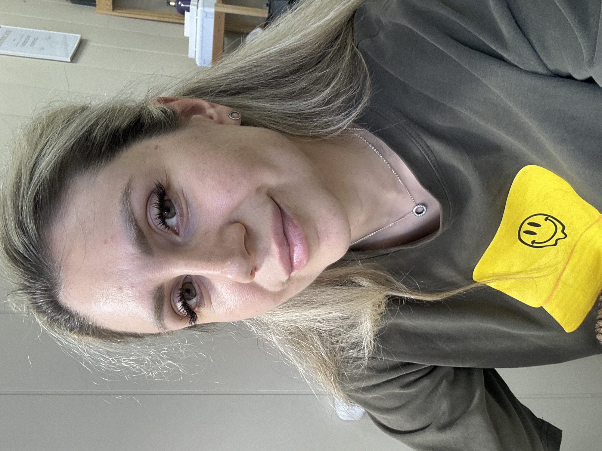 The media file is a selfie of a young woman with blonde hair, wearing a dark green t-shirt with a yellow smiley face graphic on it. She is wearing a silver necklace with a circular pendant. Her hair is long and slightly messy, falling around her face and shoulders. She has light makeup on, emphasizing her eyes and lips. The background appears to be an indoor setting with what looks like wooden shelves and some white objects in the upper left corner. The lighting suggests it might be daytime, but the exact time is not discernible. The woman's expression is neutral, with a slight smile. There is no discernible weather or specific location cues beyond the general indoor setting.