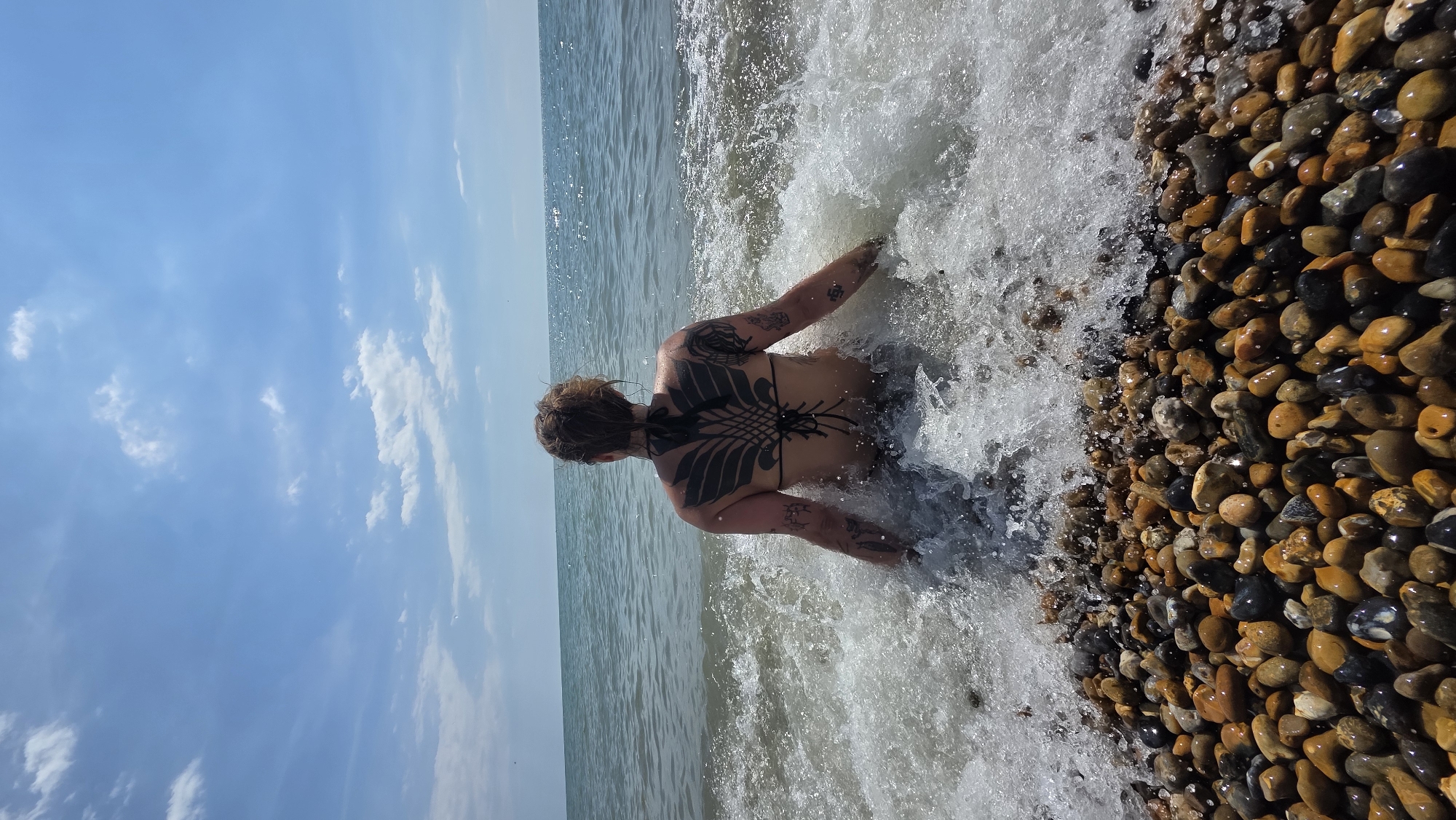 A person sits facing away from the camera, partially submerged in the ocean waves on a pebble beach. The individual has a large, dark tattoo covering their back, resembling wings. The water is churning around them, creating splashes. The sky above is blue with scattered white clouds. The foreground is dominated by a dense collection of smooth, multicolored pebbles. This scene is located in St Leonards, United Kingdom.