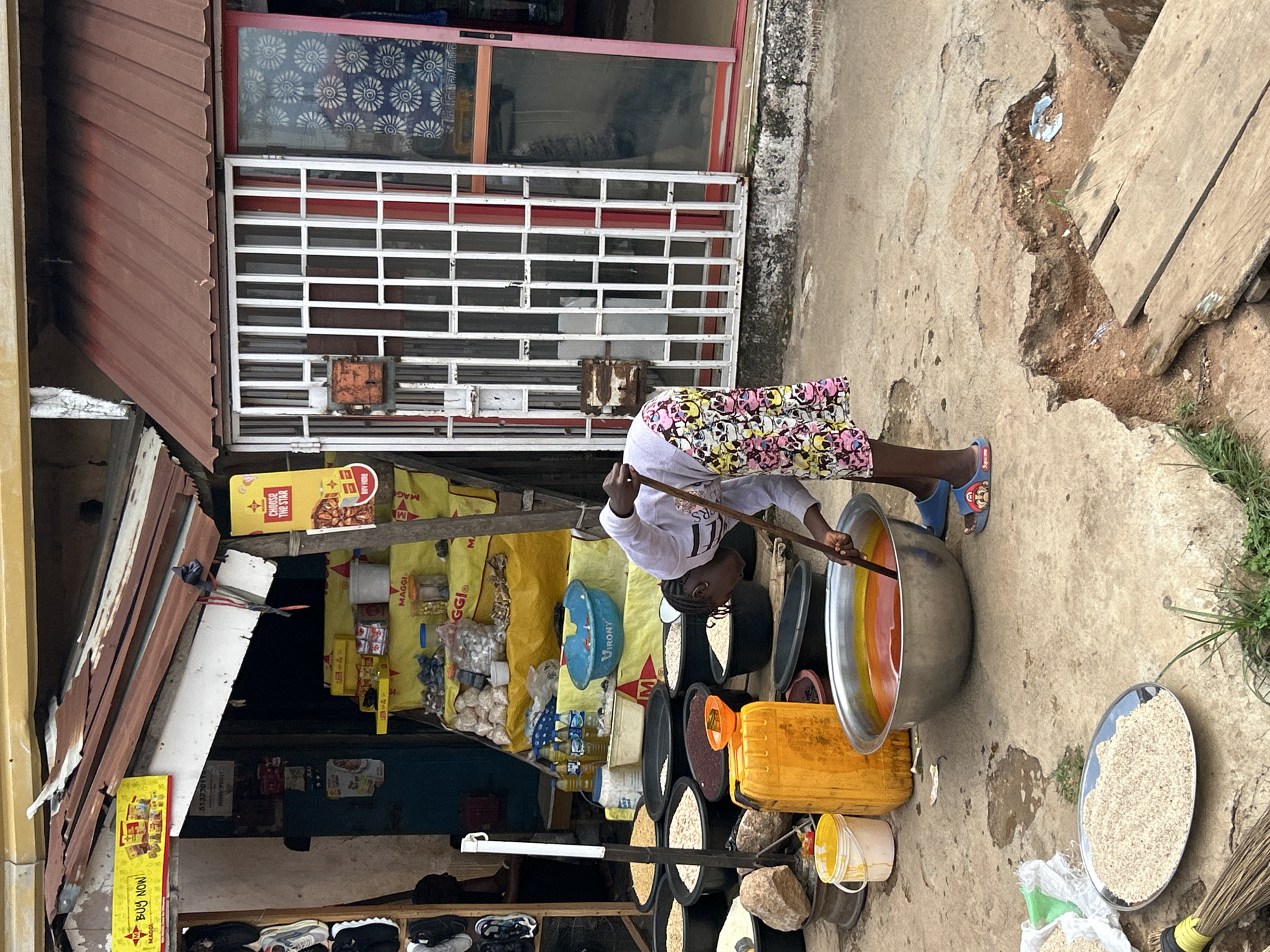 This image depicts a bustling street market scene in Jos, Nigeria, under natural daylight. The central focus is a person, likely a woman or girl, dressed in a white long-sleeved top, patterned shorts adorned with skull motifs, and blue sandals. She is actively engaged in stirring a large, shiny silver pot with a long wooden stick. The pot holds a vibrant, multi-layered liquid displaying shades of orange, yellow, and possibly red, suggesting food or drink preparation for sale.
Around the person, the ground is an uneven, cracked concrete surface with patches of dirt and a wooden plank. Various items are arranged for sale: several black buckets and containers are filled with different dry goods, such as grains or powders. A yellow jerry can is positioned near the large pot, and a blue plastic basin marked with "VIRONY" is visible amidst other goods. To the right, an oval metal tray is laden with a pile of light-colored granular material, possibly groundnuts or a type of flour, next to a broom.
The background features multiple market stalls and storefronts. One stall is heavily stocked with yellow bags, many prominently displaying the brand "MAGGI," alongside other packaged items. Another shop entrance is secured by a white metal grid gate, with a red-framed window above it featuring a blue and white patterned design. Corrugated metal roofing covers parts of these structures, indicating a semi-permanent market setup.
Visible text includes multiple instances of "BUY NOW," "MAGGI," "VIRONY," "CHOOSE THE STAR," and "BUY HERE." Other less clear texts include "5132301" and "MONS POUCT." The scene conveys the lively atmosphere of a typical local marketplace.