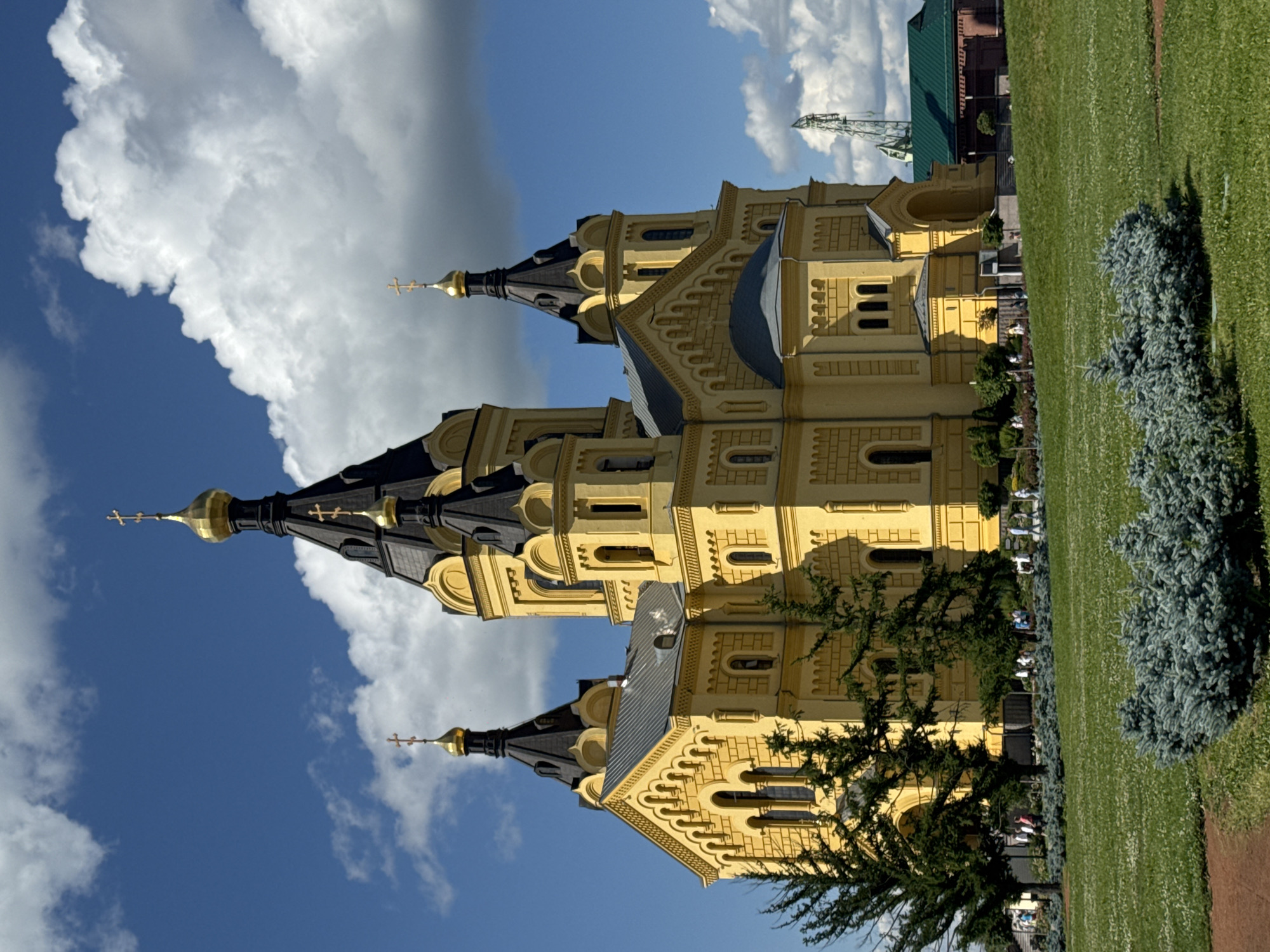 This image depicts the Alexander Nevsky Cathedral in Nizhny Novgorod, Russia, bathed in sunlight under a partly cloudy sky. The striking yellow facade of the cathedral, with its multiple black onion domes topped with golden crosses, dominates the frame. The architectural style is distinctly Russian Orthodox. In the foreground, a lush green lawn is dotted with small white flowers, and a cluster of low-lying, silvery-green evergreen bushes occupies the lower right corner. Several mature evergreen trees are positioned to the left and center, partially obscuring the cathedral's lower levels. In the middle ground, a pathway lined with more landscaping and scattered figures of people walking are visible. Beyond the cathedral, a section of a green roof and a tall, green structure, possibly part of a industrial crane or tower, can be seen against the blue sky with puffy white clouds. The overall scene conveys a sense of architectural grandeur within a well-maintained public space on a clear day.