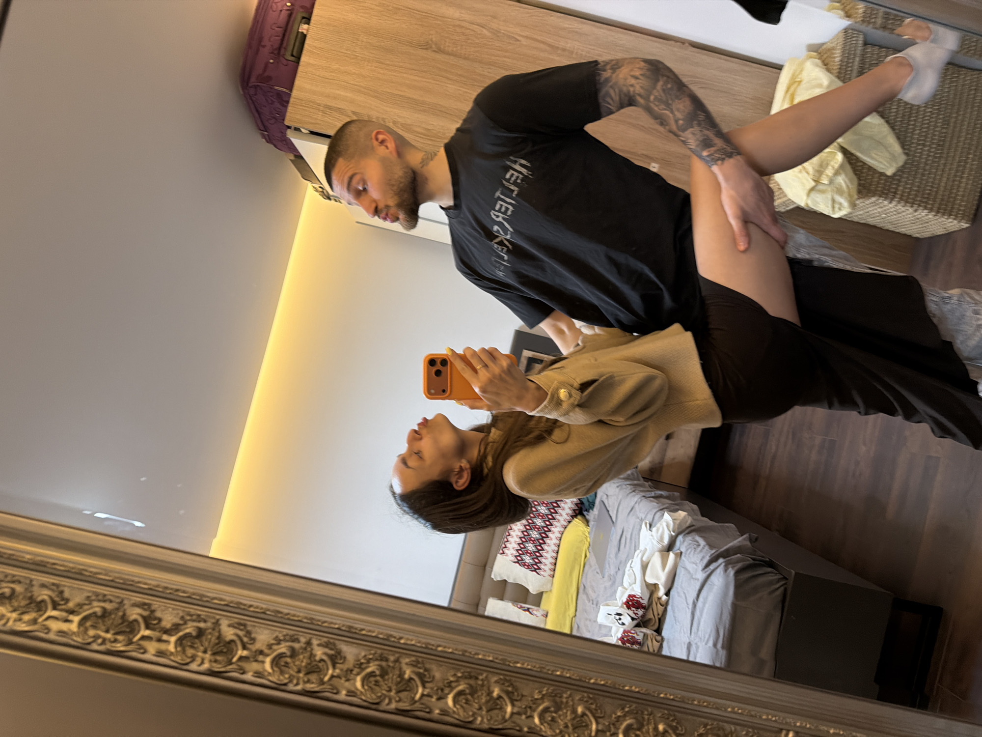 A woman holds up an orange smartphone in front of a mirror to photograph herself with a man, as reflected in the mirror. The mirror is ornately framed in silver. The man is fair-skinned with short hair, a beard, and tattoos on his left arm. He is wearing a black T-shirt. The woman has long brown hair and wears a tan jacket. They are in a bedroom with a bed, a dresser, and a laundry basket. There is also a rolling suitcase. The location is most likely Sofia, Bulgaria.