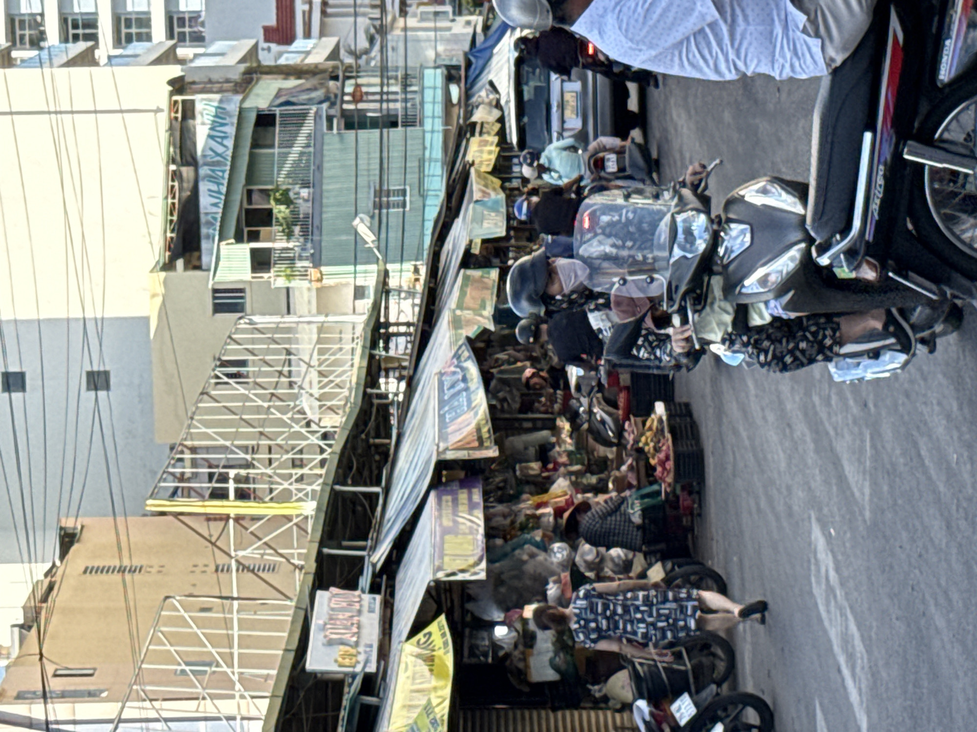 The image captures a street scene in Nha Trang, Vietnam, with a market area on the left and buildings in the background. In the foreground, a person wearing a patterned dress and black shoes walks away from the camera. To their right, several motorcycles are parked. Further back, market stalls are visible, displaying various goods. People on motorcycles are seen in the middle ground, some wearing helmets and face masks, indicating traffic and daily life. The buildings in the background are a mix of modern and older architecture, with visible signs and awnings over the market stalls. The overall impression is of a bustling urban environment, likely during daylight hours.