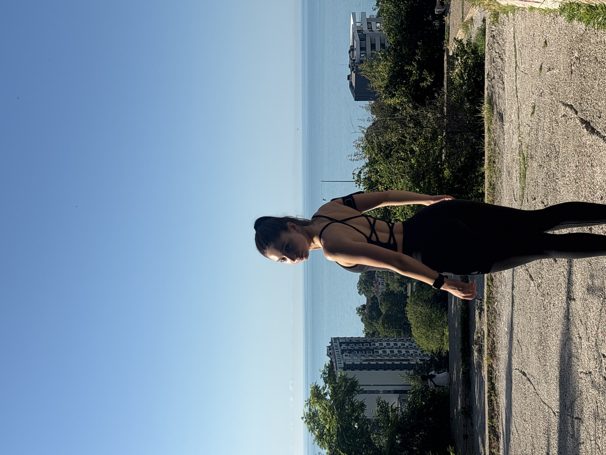 A woman wearing a black sports bra and black leggings stands in a park, looking over her shoulder towards the sea. She has her hair pulled back in a ponytail and is wearing a smartwatch on her left wrist. Behind her, the sea stretches to the horizon, meeting a clear blue sky. In the background, several buildings and lush green trees are visible, characteristic of the city of Varna, Bulgaria, particularly along its coastline. The ground in the foreground is paved with rough, textured concrete, with some cracks and patches of dry grass growing through. The lighting suggests it is a sunny day, casting shadows on the ground. A person in a white shirt is sitting on a ledge in the middle ground, near a tall building.
