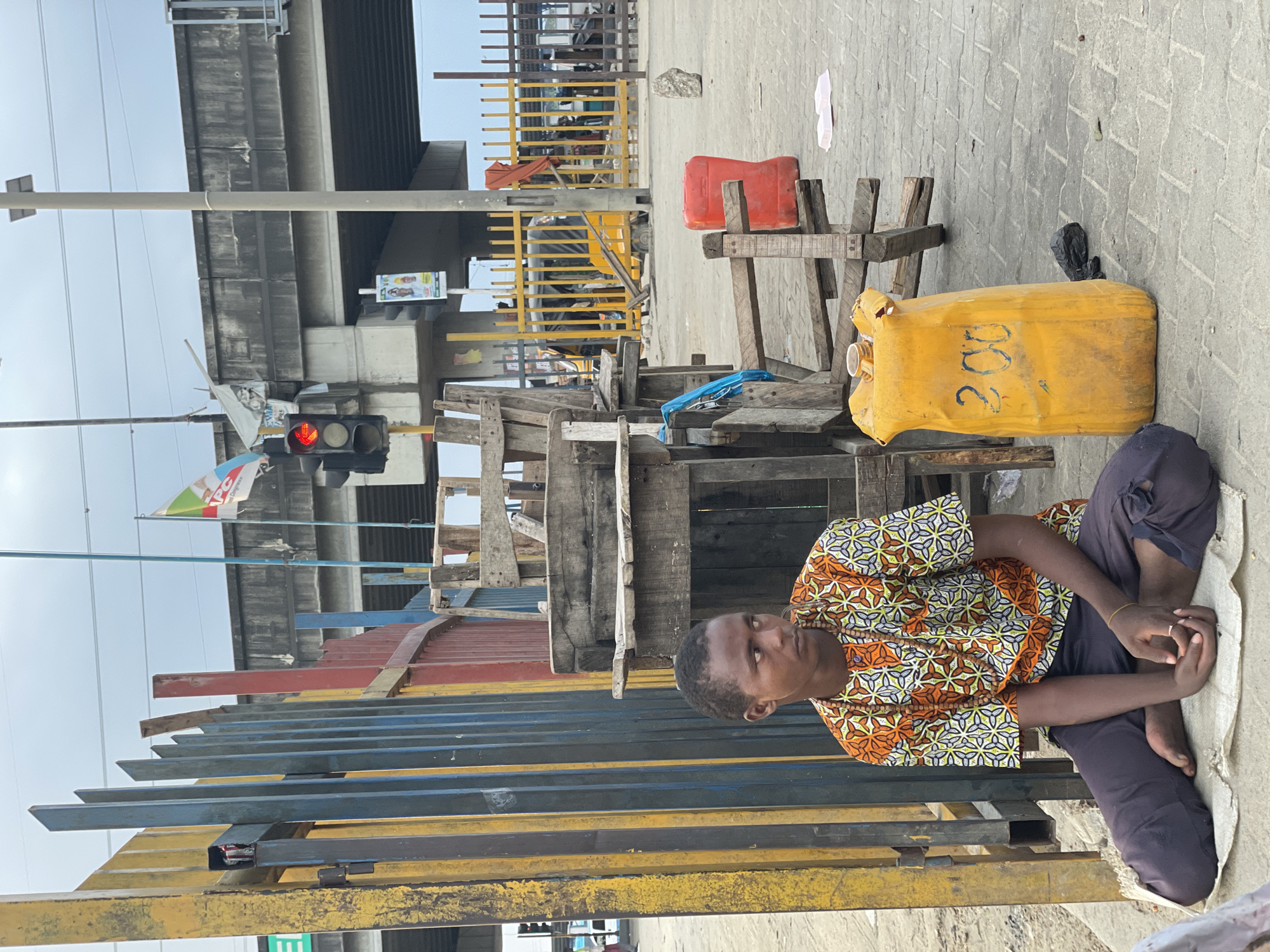 A young man sits cross-legged on a mat on a paved city street in Lagos, Nigeria. He wears a colorful, patterned short-sleeved shirt and dark pants. Beside him is a large yellow plastic container with "200" written on it in black marker. In the background, a multi-level overpass looms above. A traffic light, showing red, is visible near the overpass. A colorful metal fence runs along the left side of the image, with a wooden structure behind the young man. The overall environment suggests an urban setting, possibly near a busy road or construction area.