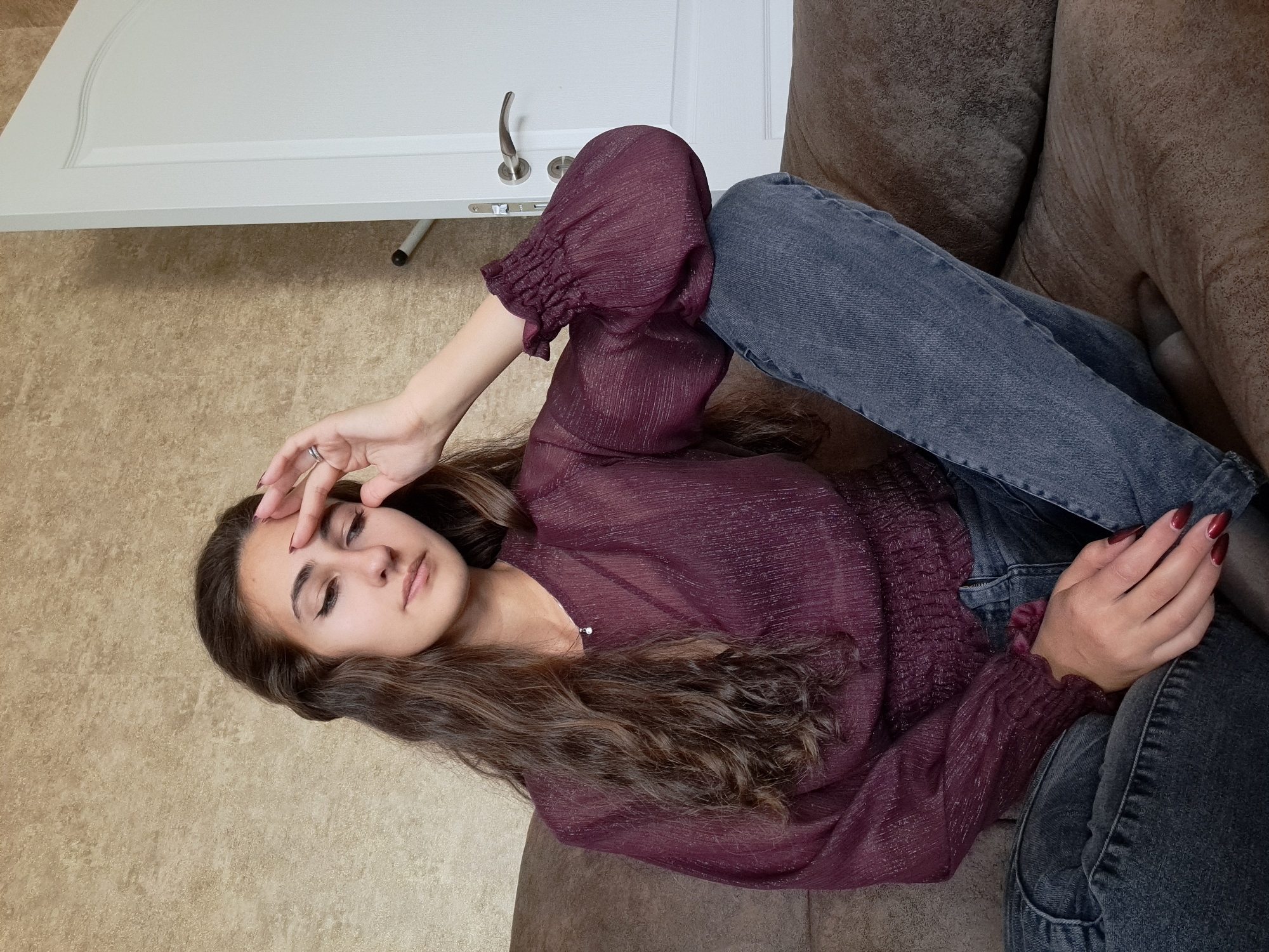 A young woman with long, wavy brown hair is reclining on a brown sofa. She is wearing a sheer, long-sleeved burgundy blouse and gray jeans. Her left hand is resting on her forehead, while her right hand is clasped in her lap. The background includes a beige wall and a white door with a silver handle. There is no indication that this image was taken in Negovan, Bulgaria.