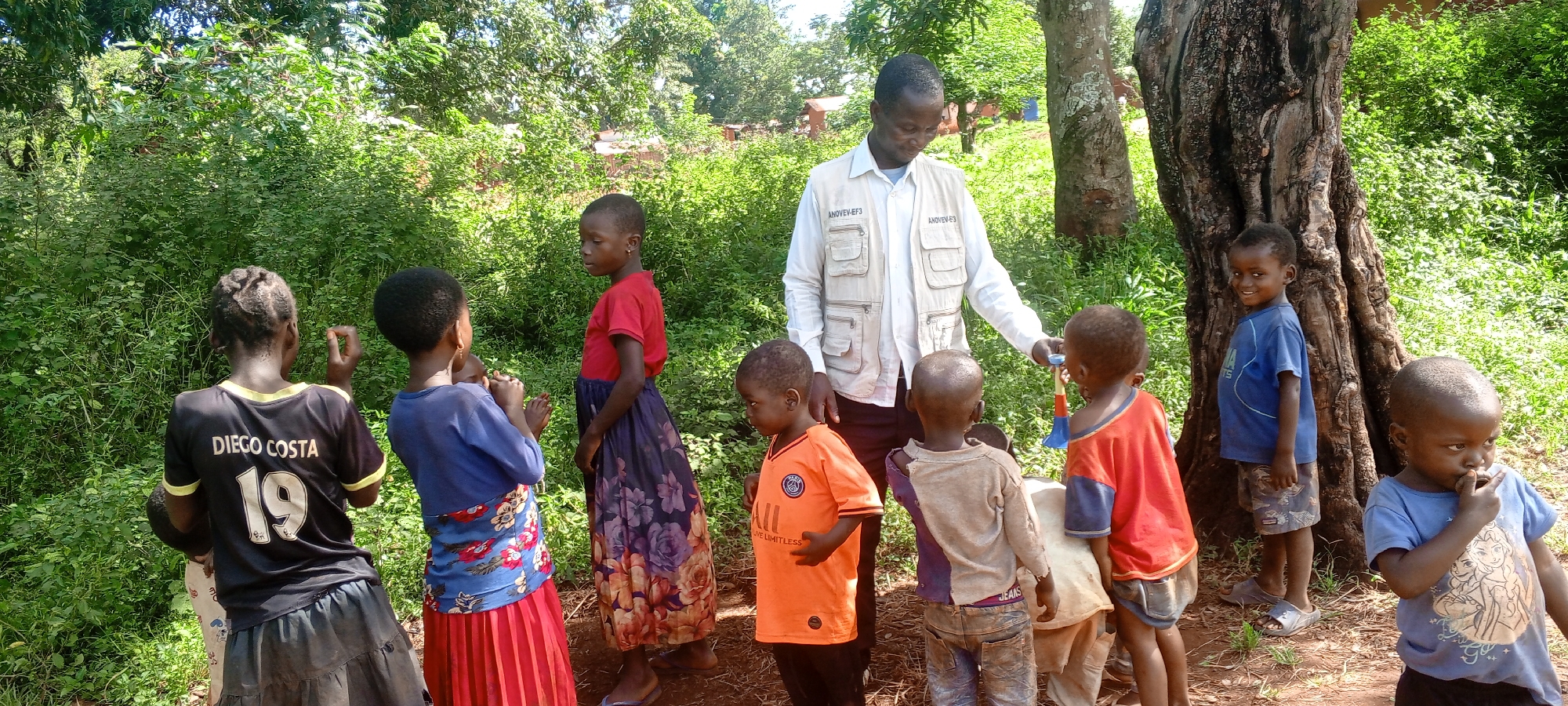The image shows a group of children gathered around an adult male in a forested area. The location is likely Ngalula, Democratic Republic of the Congo, given the vegetation and demographics.
The adult male is wearing a white button-down shirt and a light-colored vest with writing on it. He is holding a small object, possibly a toy or musical instrument, in his hand.
The children, who appear to be of African descent, are of varying ages. They are wearing a mix of clothing, including t-shirts, skirts, and dresses. One child is wearing a black soccer jersey with "DIEGO COSTA 19" on the back.
The environment consists of dense green foliage, trees, and exposed red soil. In the background, there are glimpses of what may be rudimentary structures or dwellings. The lighting suggests it is daytime.
