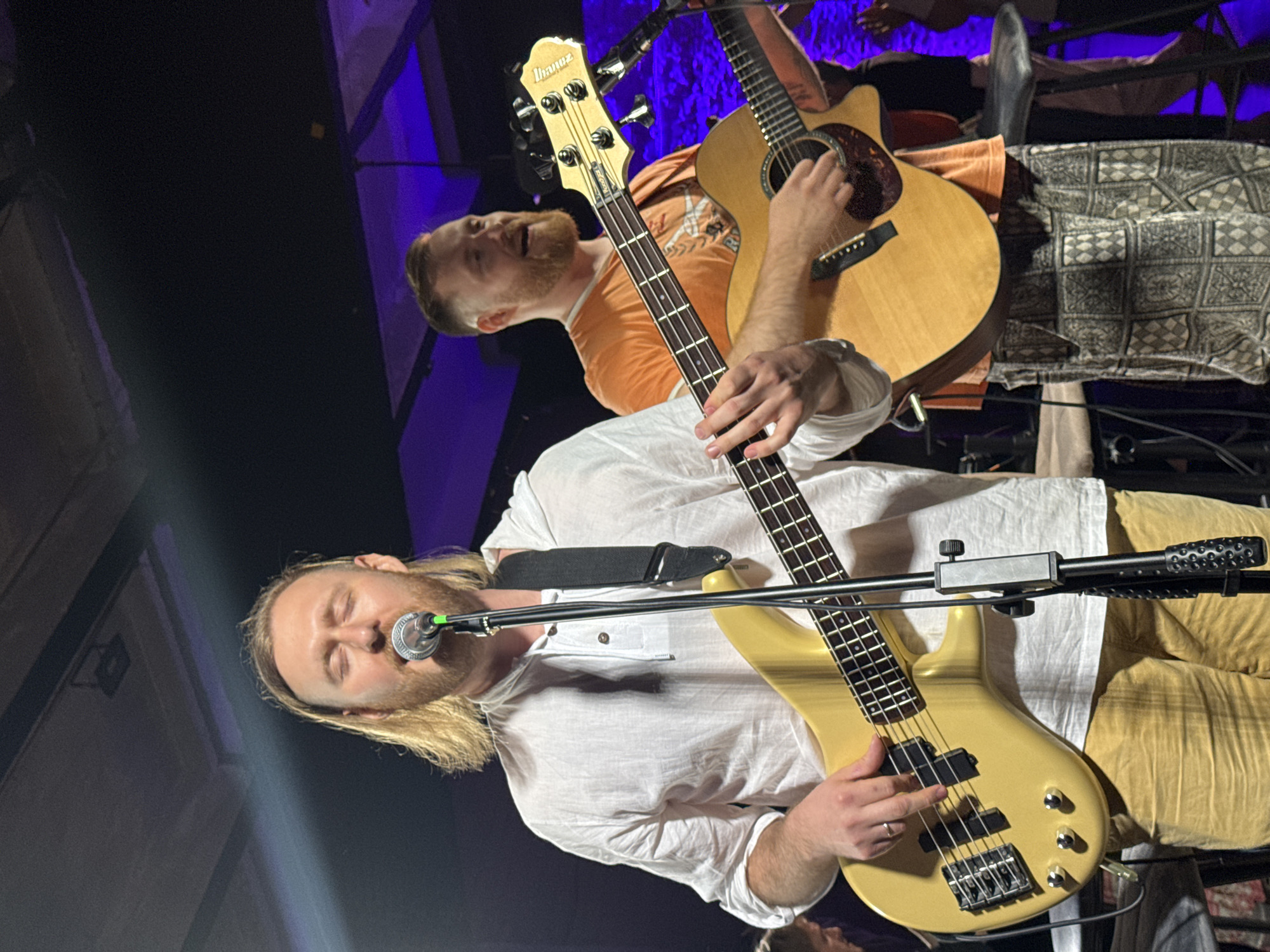 The image shows two men playing musical instruments. The man in the foreground is singing into a microphone while playing a gold-colored bass guitar. He has long blonde hair and is wearing a white shirt and tan pants. Behind him, another man is playing an acoustic guitar. He has a beard and is wearing an orange shirt and patterned pants. The location appears to be a stage or performance area. The environment is dark, with some blue lighting in the background.