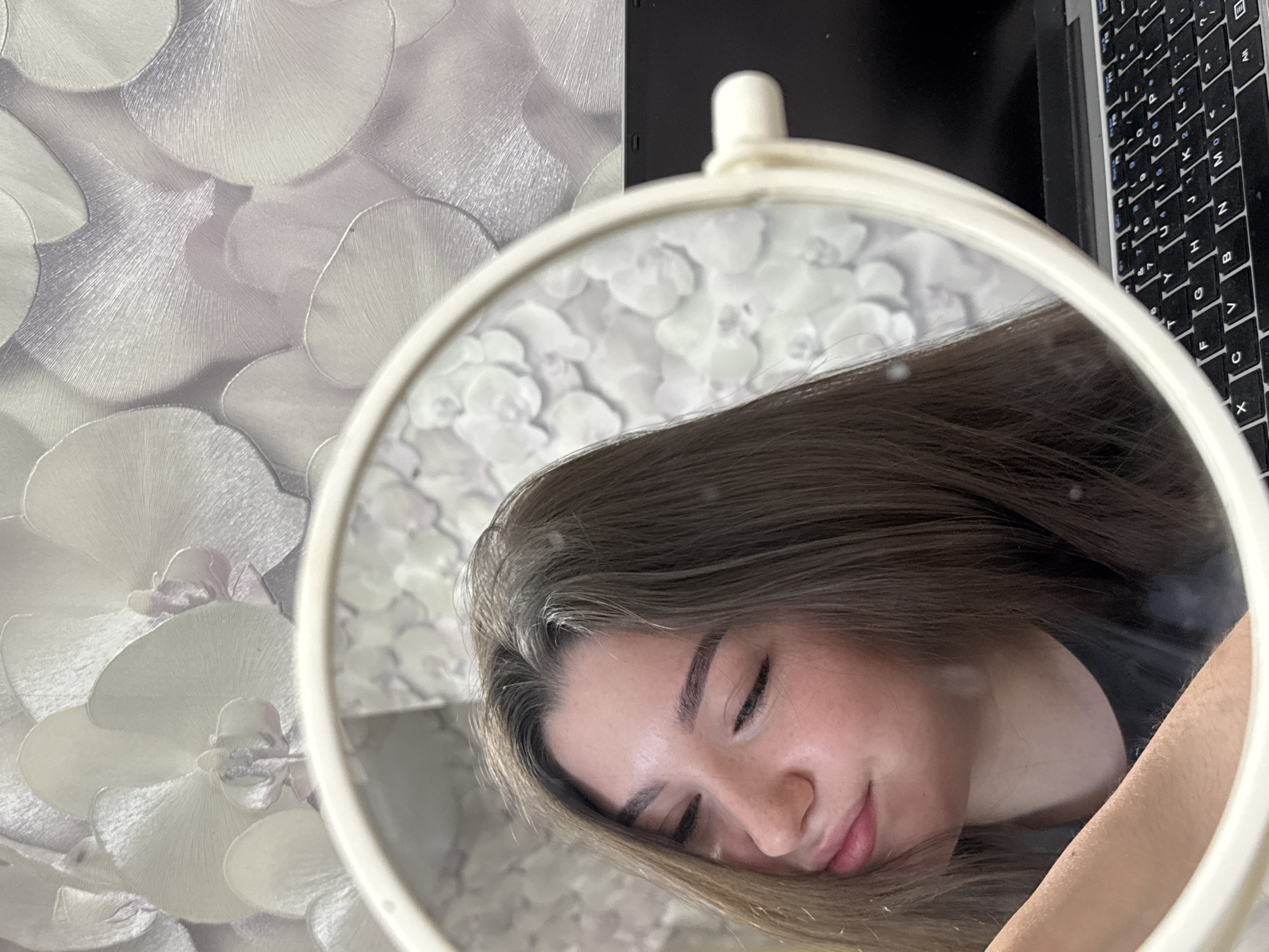 Here is a detailed description of the image:
**People:** The image prominently features a young woman, likely in her late teens or early twenties. Her eyes are closed, suggesting she is resting or sleeping. She has fair skin, defined eyebrows, and long brown hair.
**Subjects:** The primary subject is the woman's reflection in a round, white-framed mirror. The mirror's handle is visible at the top.
**Objects:** In addition to the woman and the mirror, other objects are visible. The wallpaper in the background has a floral pattern of large, white petals. A black laptop with a keyboard is partially visible behind and to the right of the mirror.
**Actions and Interactions:** The woman is passively resting, with her head slightly tilted. The mirror reflects her image, providing a view of her face from a slightly different angle.
**Environment:** The setting appears to be indoors, likely a bedroom or living space, based on the wallpaper and the presence of a laptop.
**Location:** Although the image does not provide specific clues to confirm the location, it is stated that it may have been taken in Perm, Russia. However, without additional information or visual indicators, this cannot be definitively determined from the image alone.