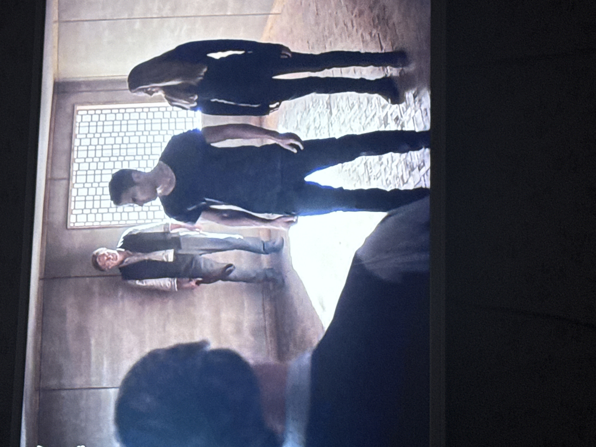 A group of three people stand inside a room with gray walls and floors. The person closest to the camera is wearing a suit. Another person is wearing a dark t-shirt and pants, while the third person is wearing all dark clothing. A square, latticed window is visible in the background. The image does not contain information to identify a location in Mytishchi, Russia.
