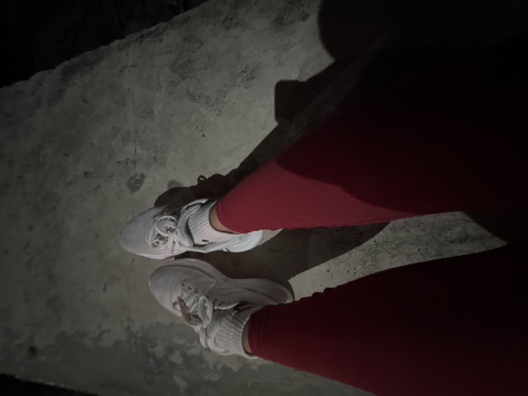 The image presents a first-person, downward perspective of a person's lower legs and feet. The individual is wearing bright red leggings or pants, white socks, and white athletic shoes with laces. They are standing on a textured, grey concrete or paved surface that appears somewhat rough and uneven. The scene is notably dark, suggesting it is nighttime or a very dimly lit environment. A localized, bright light source (likely a flash) illuminates the immediate area around the feet, casting long, distinct shadows of the legs and shoes onto the ground. No other subjects, objects, or environmental features are clearly visible in the dark background, nor is there any visible text. The activity depicted is simply standing or a momentary pause. This scene is situated in Davao City, Philippines.