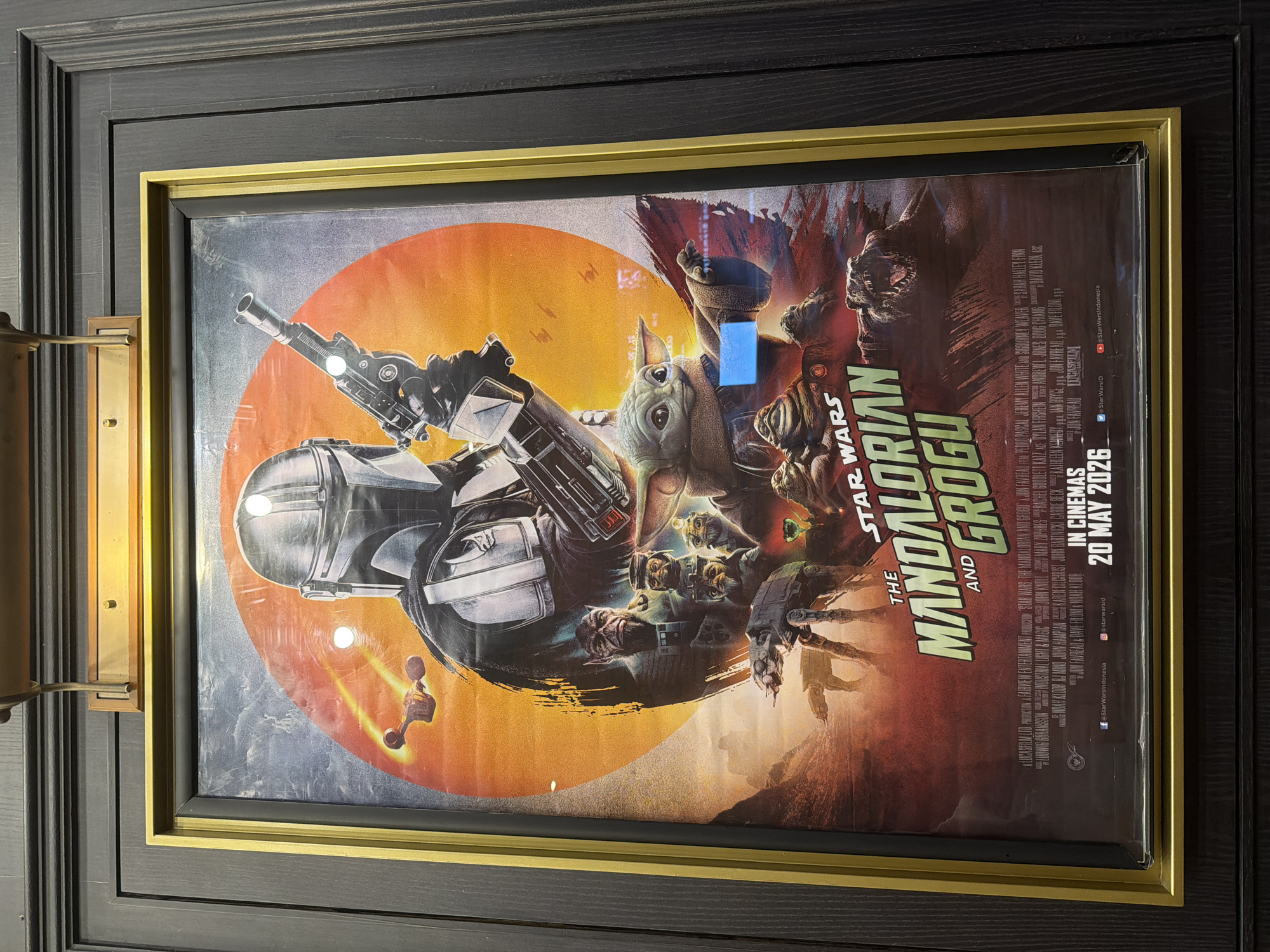 The image shows a framed movie poster for "Star Wars: The Mandalorian and Grogu", dated for release on May 20, 2026. The poster features characters from the Star Wars universe in a circular design with an orange and grey color scheme. The poster is framed with a gold inner frame and a black outer frame. The outer frame is mounted on a dark, wood-paneled wall, with a brass-colored sconce light fixture visible on the left side of the frame. The image was taken indoors. The location appears to be Unknown, Unknown.