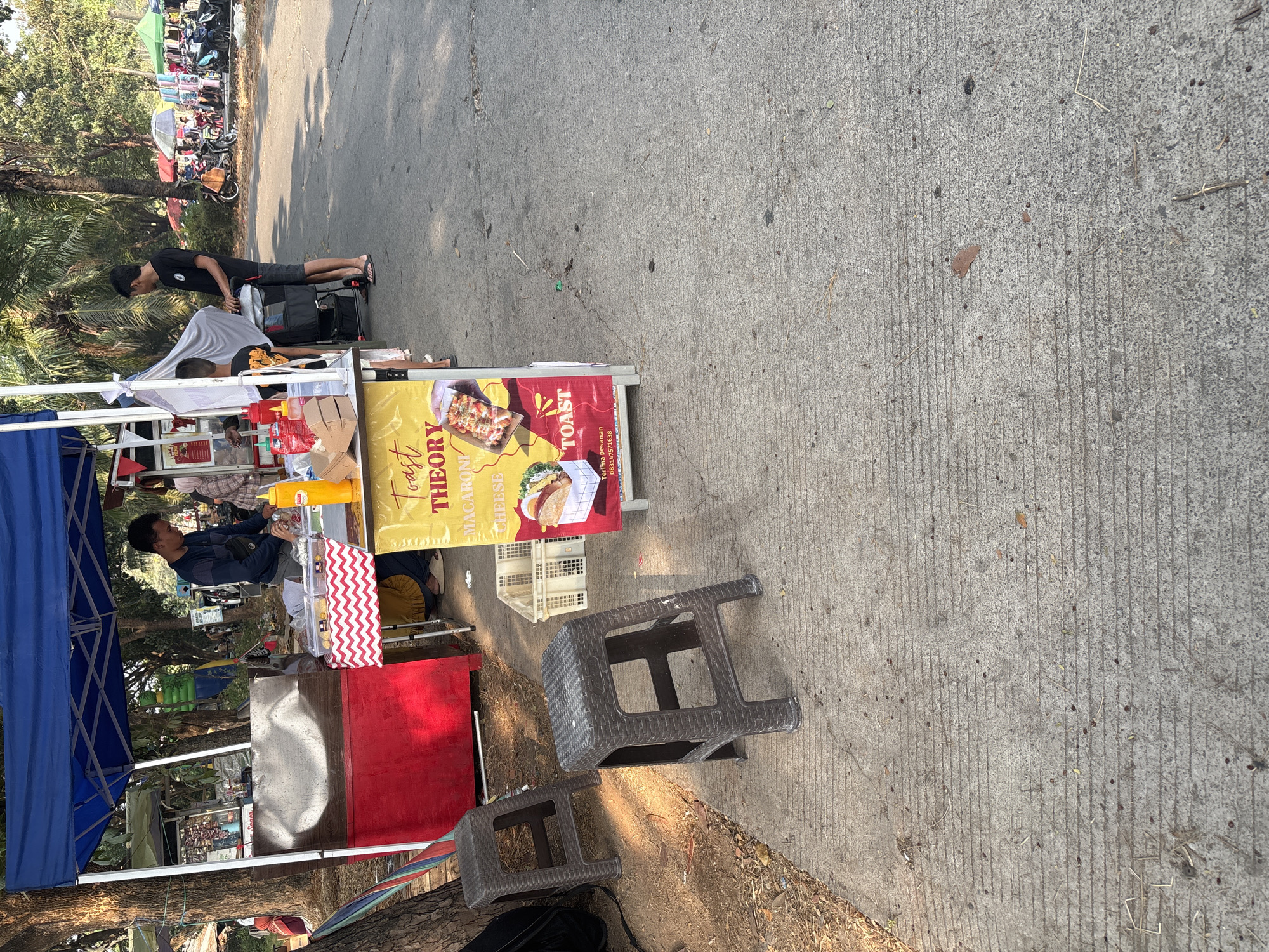 The image depicts an outdoor scene in Telagamurni, Indonesia, at a street-side food stall. A blue canopy tent is set up, offering shade over a food preparation area. Two dark brown plastic stools are positioned on a concrete walkway in front of the stall.
A young man in a dark long-sleeved shirt and shorts is standing behind the counter, actively engaged in food preparation. He is reaching towards a bright yellow bottle. Another individual, a young boy in a dark shirt with a graphic print, is standing near a stroller, facing away from the camera.
The food stall features a red base with a white and red chevron-patterned cloth draped over a table. A sign with a yellow background and red lettering advertises "TOAST THEORY MACARONI CHEESE," with images of food items. Several food containers and bottles are visible on the counter.
In the background, a busy street scene unfolds, with various stalls, colorful umbrellas, and parked motorcycles. Trees and foliage surround the area, suggesting a park or open public space. The ground is paved with concrete, showing some wear and tear. The lighting indicates it is daytime, with sunlight casting shadows.