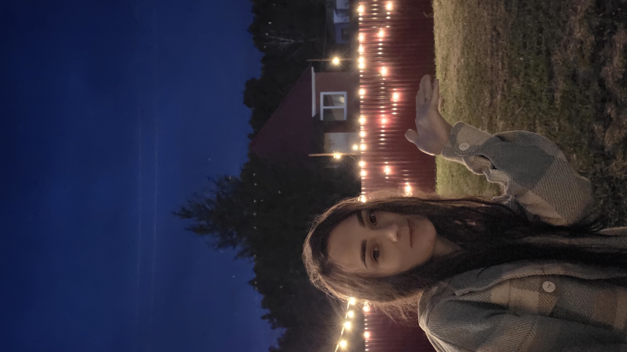 A medium shot captures a young woman with long, dark hair and light-colored eyes. She is wearing a plaid jacket in shades of blue and beige. The woman is looking towards the camera with a slight smile and her right hand is raised, palm open, as if presenting something. Behind her, a red fence is illuminated by a string of warm-toned lights. In the background, a small, red-roofed house is visible, surrounded by trees. The sky is a deep blue, suggesting it is twilight or night. The scene appears to be outdoors in a backyard setting.