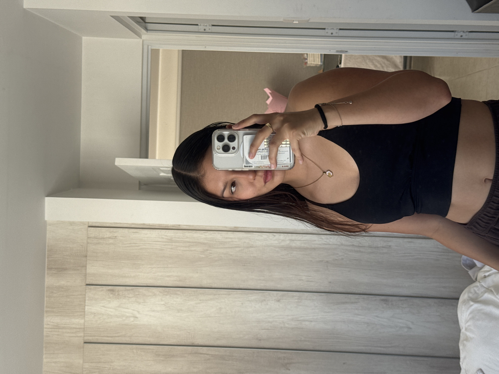 A young woman is captured in a mirror selfie, holding a smartphone with a clear case. She has dark, straight hair, parted down the middle, and is looking directly at her reflection in the phone. Her expression is neutral. She wears a black sports bra or crop top, a gold necklace with a light-colored pendant, a gold ring on her left hand, and bracelets on her left wrist, including one with small beads and another with a chain. Her midriff is visible.
The scene is indoors, likely a room in San Francisco Ocotlán, Mexico. The background consists of light-colored walls and a light-toned wooden door or cabinet on the left side of the frame. A white shelf or structure is visible above the wooden element. To the right, another section of the room is reflected in the mirror, showing light-colored walls and a potential doorway or opening in the distance. Part of a white sheet or blanket is visible in the bottom right corner.
On the back of her phone, through the clear case, partially obscured text including "sm.qsilbu" and what appears to be labels or a barcode are visible. Some faint, illegible text is also reflected on the phone screen itself. The time of day appears to be daytime, given the bright, even lighting.