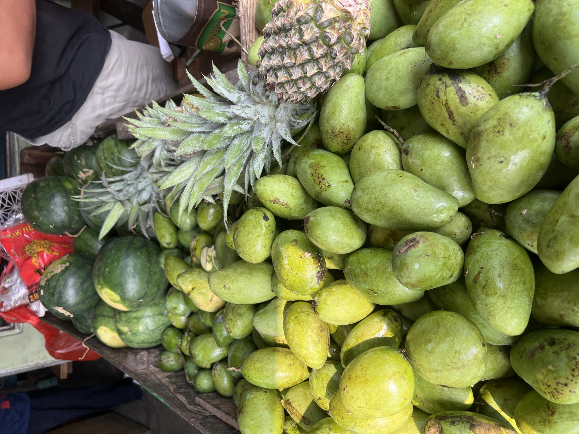 In Citeureup, Indonesia, a bustling fruit stand displays a vibrant assortment of produce. The foreground is dominated by a large pile of green mangoes, their skins glistening with a light sheen. Behind the mangoes, several pineapples with spiky green fronds are visible, some nestled among the mangoes. Further back, a selection of watermelons, their dark green rinds providing a contrast to the lighter fruits, are piled high in a wooden crate. Red plastic bags filled with an unidentifiable item are stacked behind the watermelons. A person, partially visible from the waist down, stands behind the fruit, wearing a black t-shirt and light-colored shorts, their presence suggesting they are tending to the stand. The environment appears to be outdoors, possibly a market, with the overall impression being one of abundance and fresh produce.
