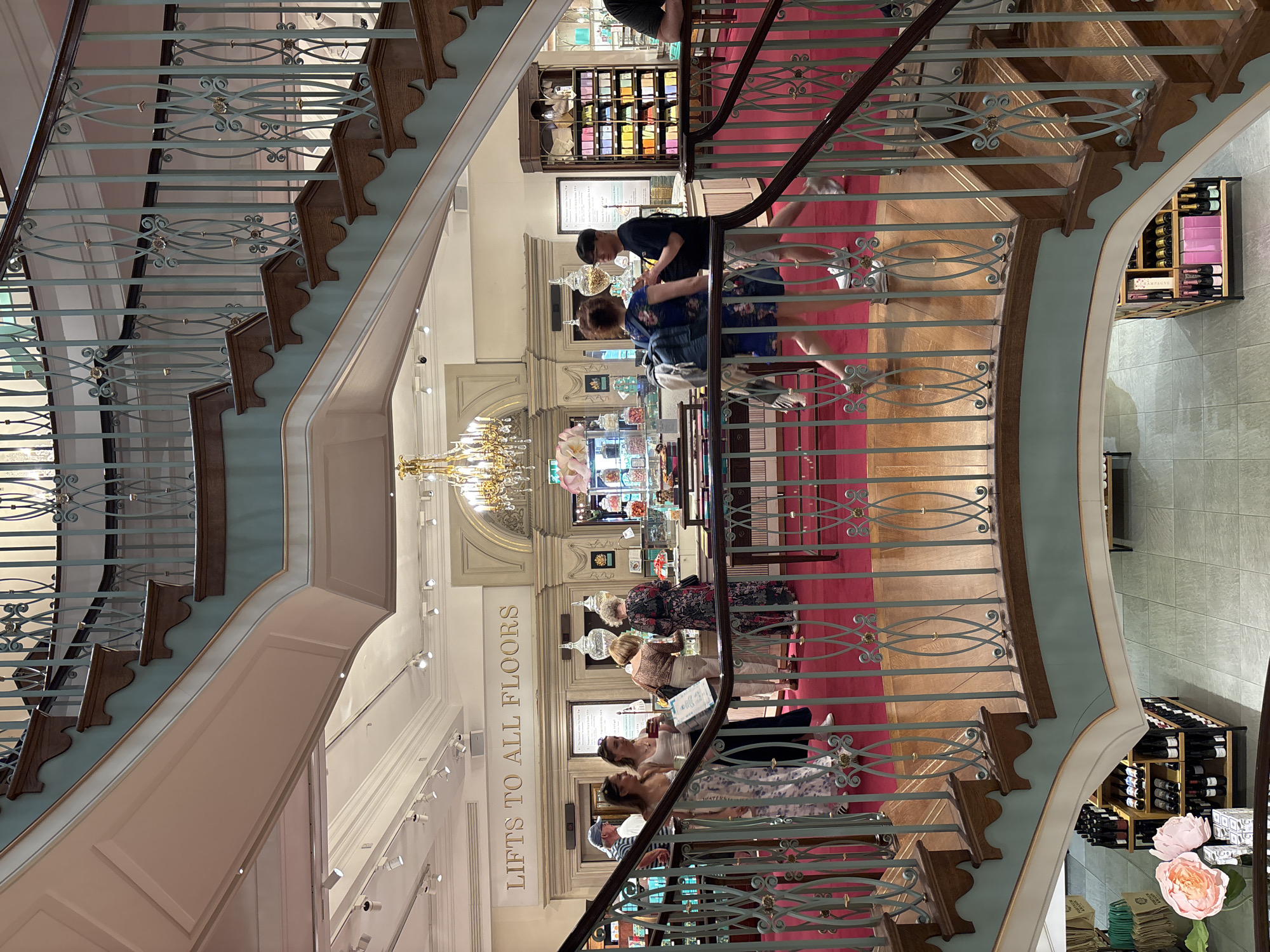 The image depicts an interior view of Fortnum & Mason in London, United Kingdom, taken from an elevated position on a staircase. The staircase features ornate light-blue railings with decorative gold accents and dark wood steps. The walls are painted in a light pink hue.
Several customers are visible browsing various departments. On the ground floor, a group of individuals is looking at a display case, and another customer is being assisted by a staff member wearing a black shirt and a face mask. A sign reading "LIFTS TO ALL FLOORS" is displayed on the wall. A chandelier is also visible on the ground floor.
Merchandise shelves, including wine, candy and tea, are placed on various floors within the building. The floors have a red carpet, and the floors are made of wood.
The image captures the upscale shopping environment and the architectural details characteristic of Fortnum & Mason.