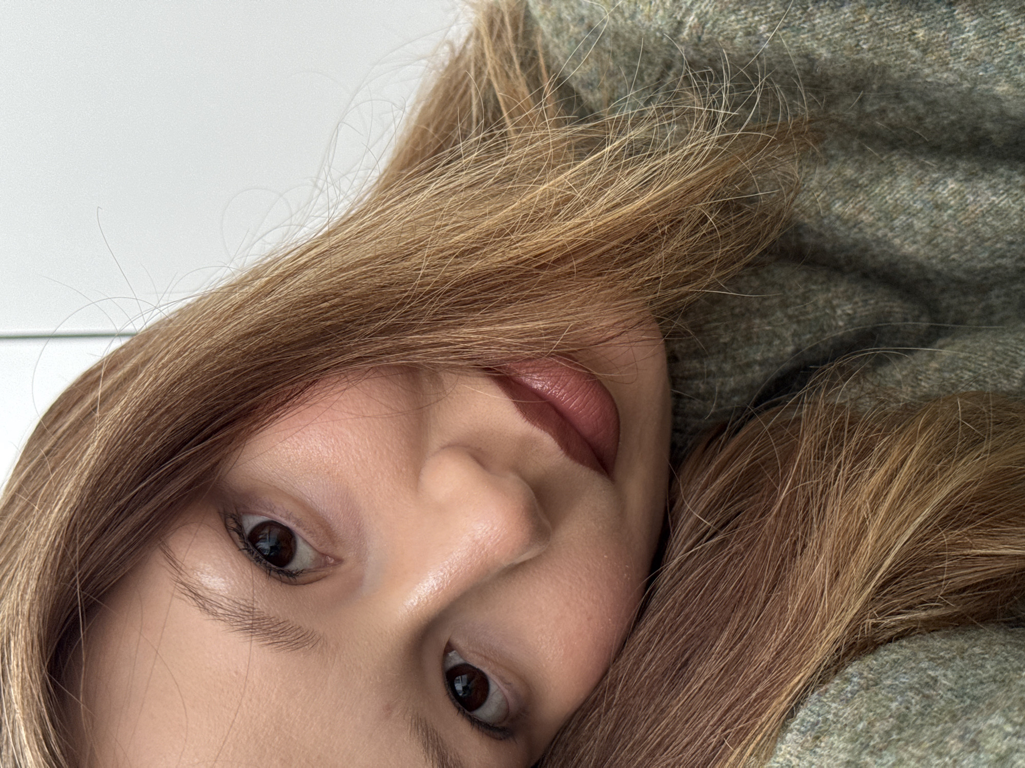 A close-up, slightly angled, indoor photograph depicts a woman's face and upper torso. She has fair skin, brown eyes, light brown hair, and is wearing lipstick. Her hair is loosely styled and falls around her face and shoulders. She is wearing a textured green sweater. The background is predominantly white, suggesting a plain wall. No location in the city is provided.