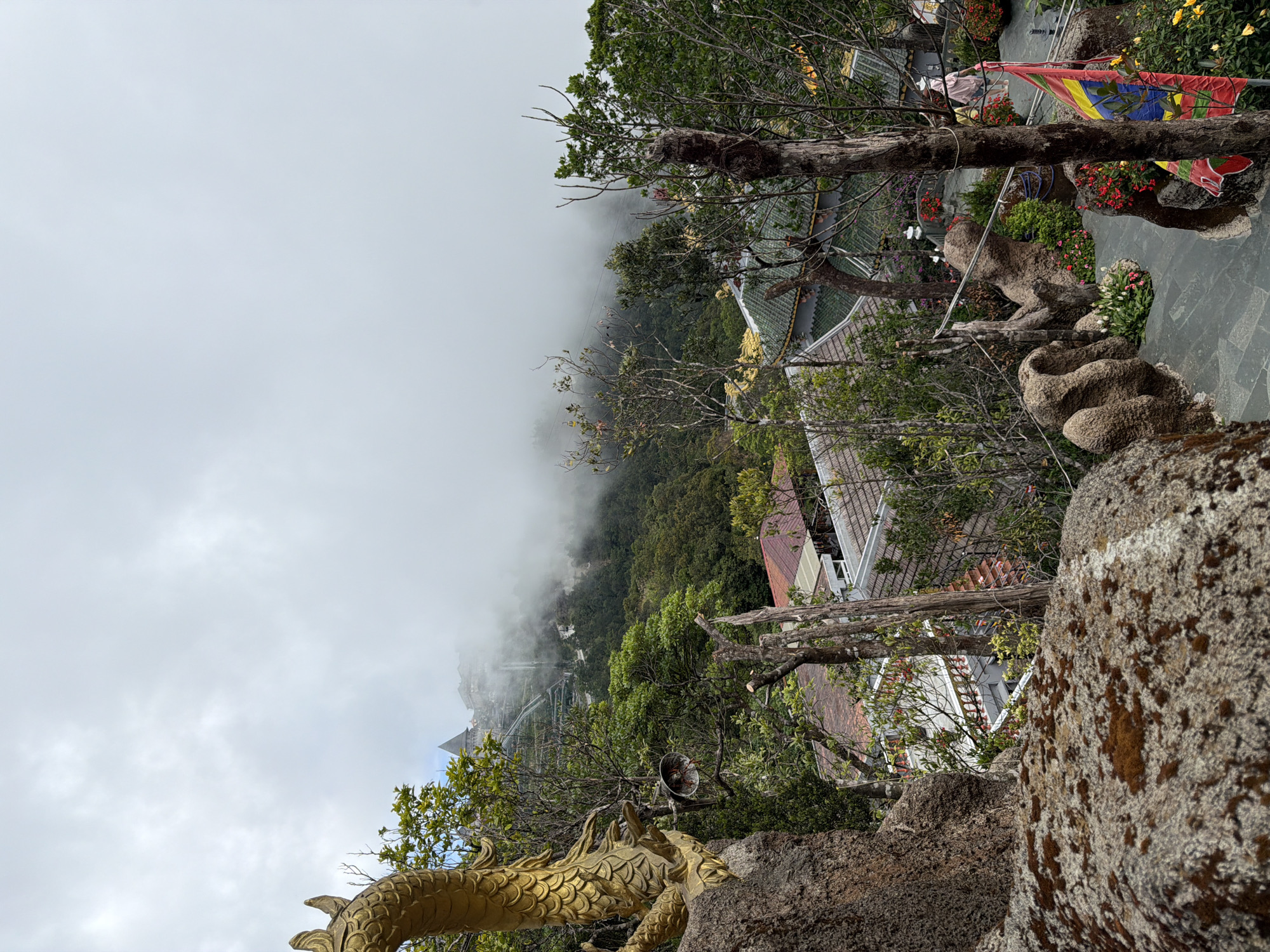 The image depicts a scenic view from a mountainous area, likely Ba Na Hills in Đà Nẵng, Vietnam. In the foreground, a large, moss-covered rock formation is visible, with a golden dragon sculpture partially framing the left side of the image. Behind the rock, a tiered landscape unfolds, featuring lush green trees, traditional Vietnamese architectural elements with green tiled roofs, and stone pathways. In the middle ground, there are glimpses of buildings and a steep, winding staircase leading up the mountain. The background is characterized by dense fog and clouds, partially obscuring more distant structures and the mountain's summit. A flag with red, yellow, and blue sections is prominent on the right side of the pathway. The overall atmosphere is serene and mystical, enhanced by the misty conditions.