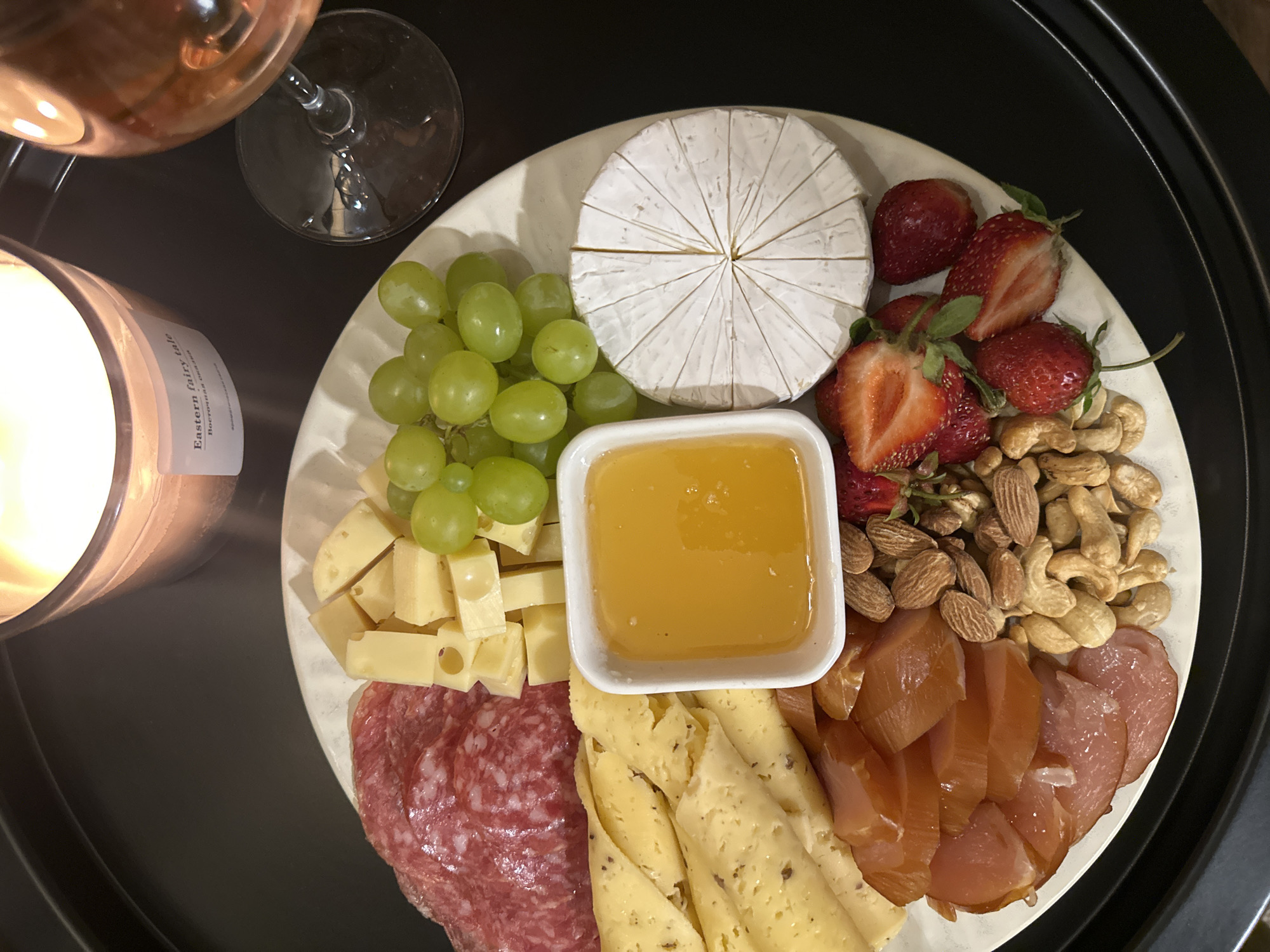 A top-down shot captures a circular white plate filled with a charcuterie assortment. The plate rests on a dark, textured tray. From the top left, the arrangement includes thin slices of red salami, followed by cubes of pale yellow cheese, and a cluster of green grapes. A small white dish containing amber-colored honey is positioned in the center. To the right of the honey is a round of white cheese, scored into wedges. Below the cheese cubes are rolled slices of a lighter yellow cheese speckled with herbs. Beneath the honey and to the left are thin slices of cured meat. The bottom of the plate features a mix of almonds and cashews, alongside sliced strawberries and whole strawberries. A lit candle with a label reading "Eastern fairy tale" sits in the upper right quadrant, and a glass of rosé wine is partially visible in the upper right corner. The overall scene evokes a sense of a cozy gathering or a personal indulgence. The location in Chaikovsky, Russia is not discernible from the image.