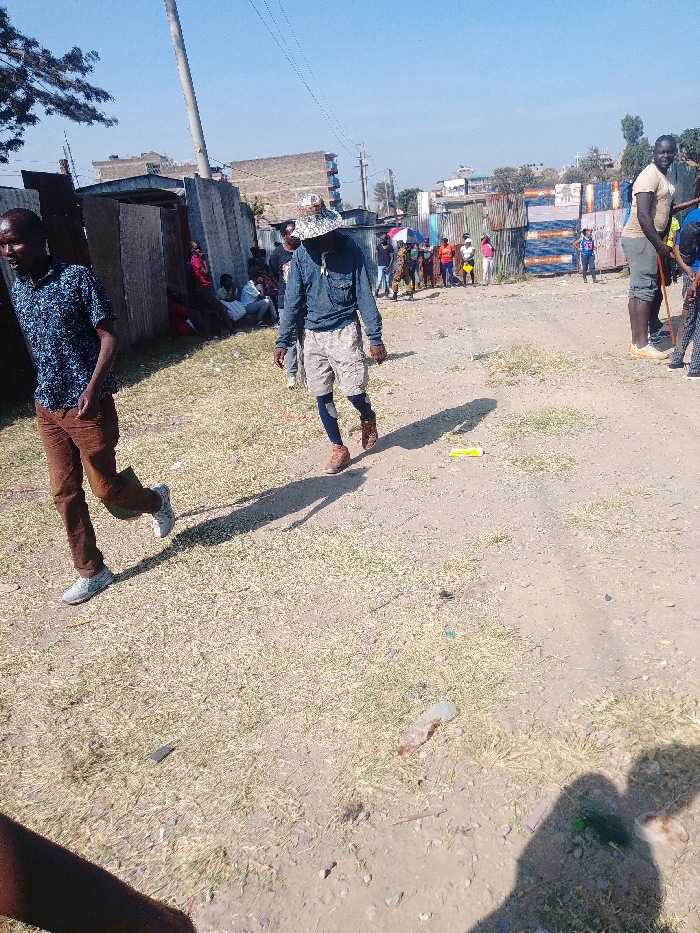 A dusty street scene features several people. One man in a blue patterned shirt and brown pants walks with another wearing a hat and denim jacket. To the right, a very tall man stands in a light tan shirt. In the background, a small crowd gathers near metal structures. The ground is a mix of dirt, grass, and scattered litter. Buildings and utility poles are visible in the distance under a bright sky.