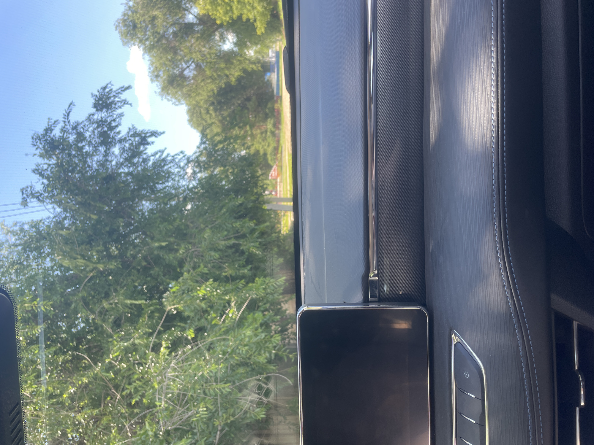 The image shows a view from inside a car dashboard looking out through the windshield. The dashboard is made of dark material with textured accents and a large screen on the left. Outside, there are trees with green leaves and a blue sky with a few clouds. A road is visible in the distance, with some buildings and greenery on either side. The scene appears to be in a suburban or rural area. The location of Линёвка, Russia is not discernible from the image.