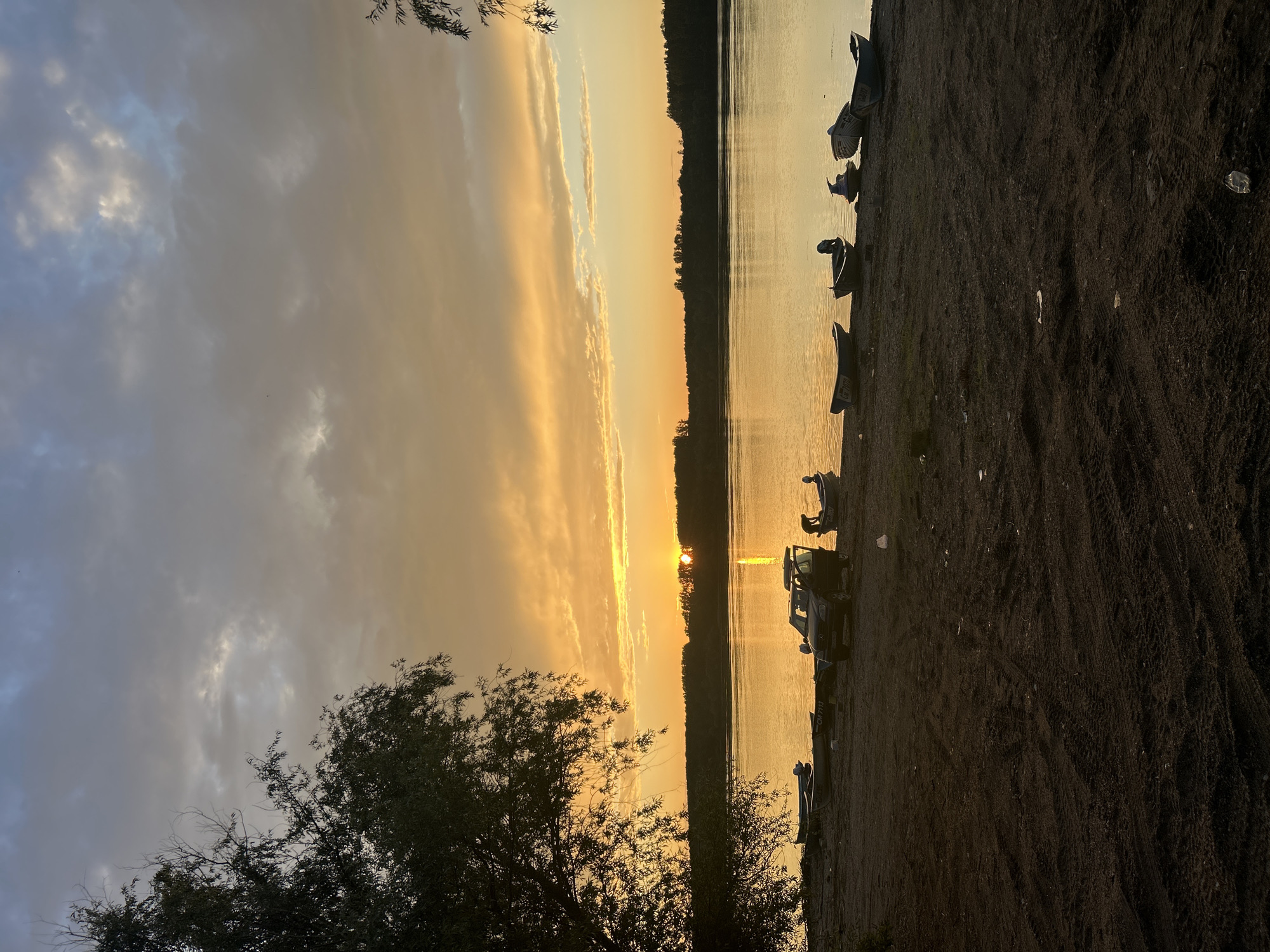 The image depicts a sunset or sunrise over the Danube River in Somovit, Bulgaria. The sky transitions from an overcast light blue and grey at the zenith to a vivid orange and yellow near the horizon, with stratocumulus clouds partially obscuring the upper atmosphere. The sun, a bright orange orb, is visible on the distant tree line, casting a golden reflection across the river's surface. A dense, dark silhouette of trees marks the far bank of the river.
In the foreground, a dark, sandy or dirt bank shows distinct tire tracks. On the left, a large deciduous tree, heavily silhouetted, dominates the scene, with its branches extending into the upper left quadrant of the frame. Multiple small boats are beached along the riverbank. From left to right, these include two small fishing boats, one bearing the registration "CM 7113". A dark-colored SUV, possibly a Mercedes-Benz, is parked facing the river with its driver's side door open. Immediately to the right of the SUV, another small boat is visible, and a human figure, silhouetted, is bent over near the rear of this boat. Further to the right, a personal watercraft resembling a jet ski is beached, followed by two additional small fishing boats, one with a visible outboard motor and another with partial registration "CM 7110". Scattered pieces of litter, including plastic bags and bottles, are visible on the sandy ground.