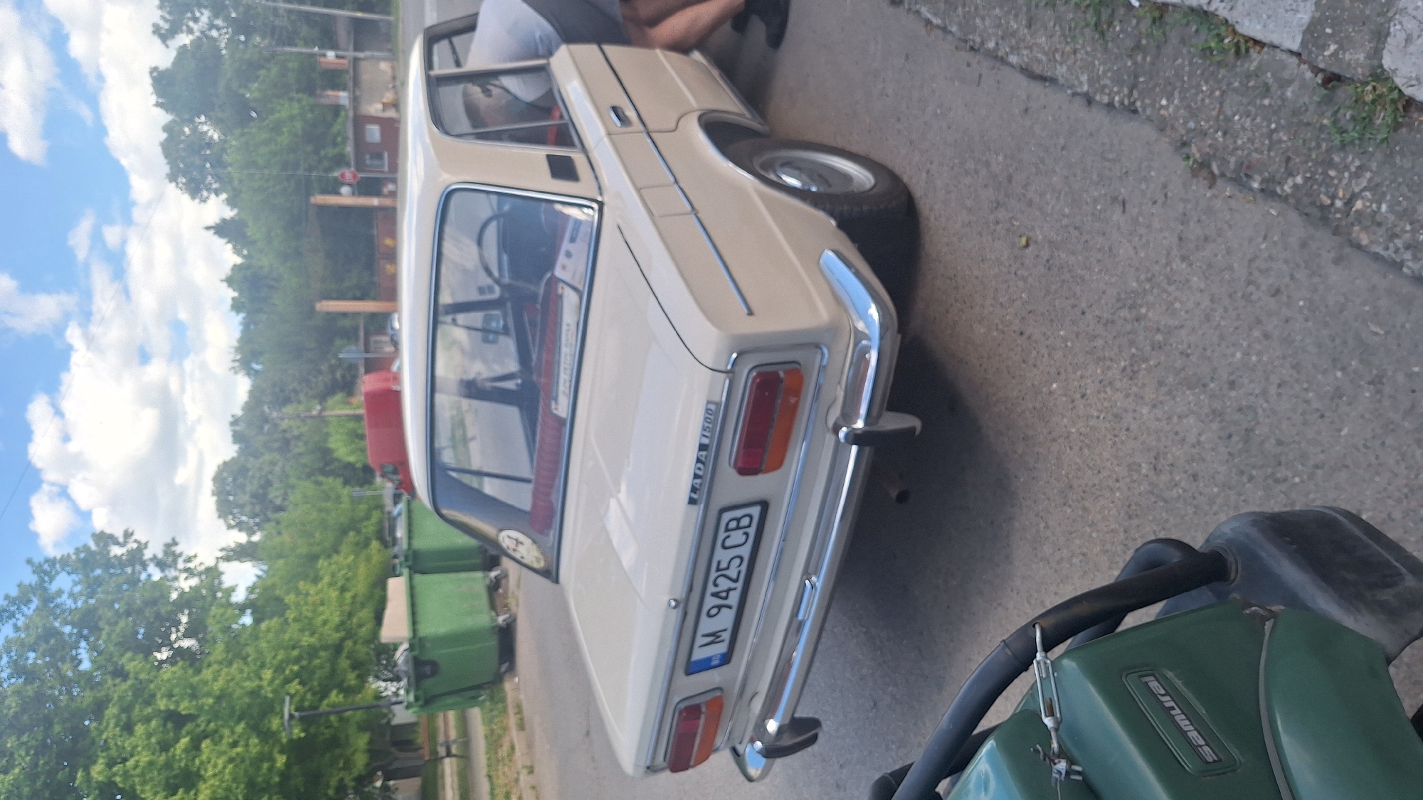 A beige Lada 1500 sedan is parked on an asphalt road in the city of Lom, Bulgaria. The car's rear is facing the camera, revealing its license plate which reads "BG M 9425 CB". To the left of the car, there are two large green dumpsters. In the background, trees and a red van are visible, along with buildings and a stop sign. The sky is partly cloudy. A person is partially visible next to the car, bending down. In the foreground, a green piece of machinery, possibly a compactor, with the word "Samurai" on it, is present.