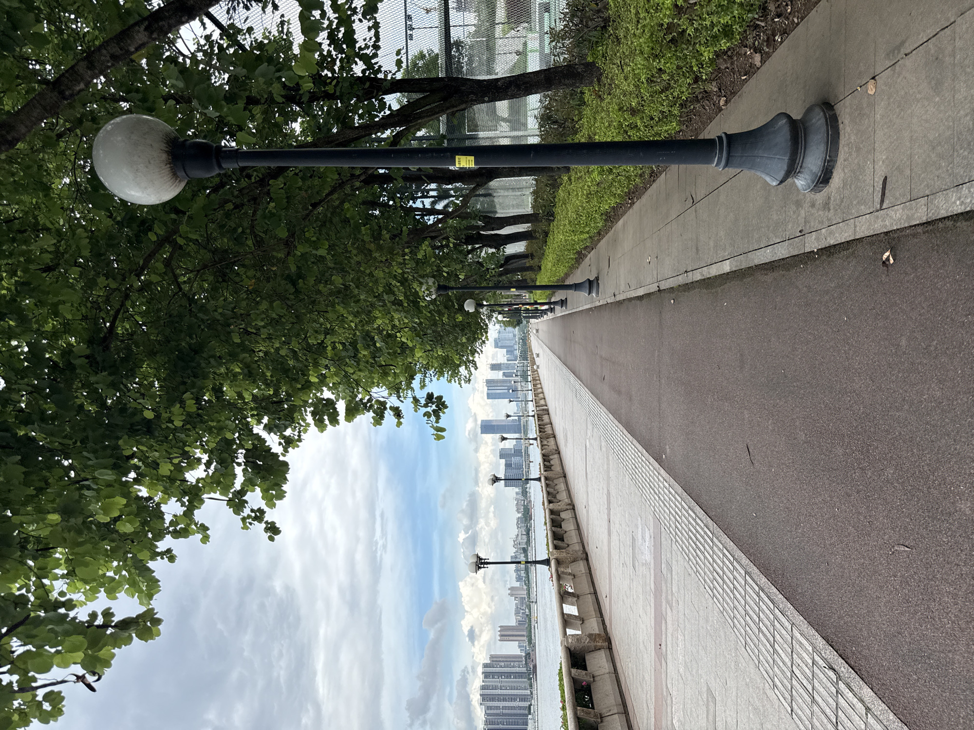 The image depicts a pathway alongside a body of water, with a city skyline in the distance. The pathway is bordered by trees and lush green bushes on one side, and a paved walkway with a textured border on the other. Several lampposts are evenly spaced along the walkway, casting a warm glow. The sky is partly cloudy, with patches of blue peeking through. The city skyline features numerous tall buildings, suggesting an urban environment.
The scene appears to be set in a park or promenade, likely in Tianhe District, China, given the context. There are no people visible in the image. The time of day is difficult to determine precisely, but the lighting suggests it could be late afternoon or early evening. The weather seems mild, with the sky indicating a possibility of light clouds.
Notable details include the decorative bases of the lampposts, the textured pattern on the walkway's edge, and the distinct architectural style of the buildings in the skyline. A small yellow sign is affixed to one of the lampposts, but the text is illegible. The overall mood of the image is peaceful and serene, inviting a stroll along the path.