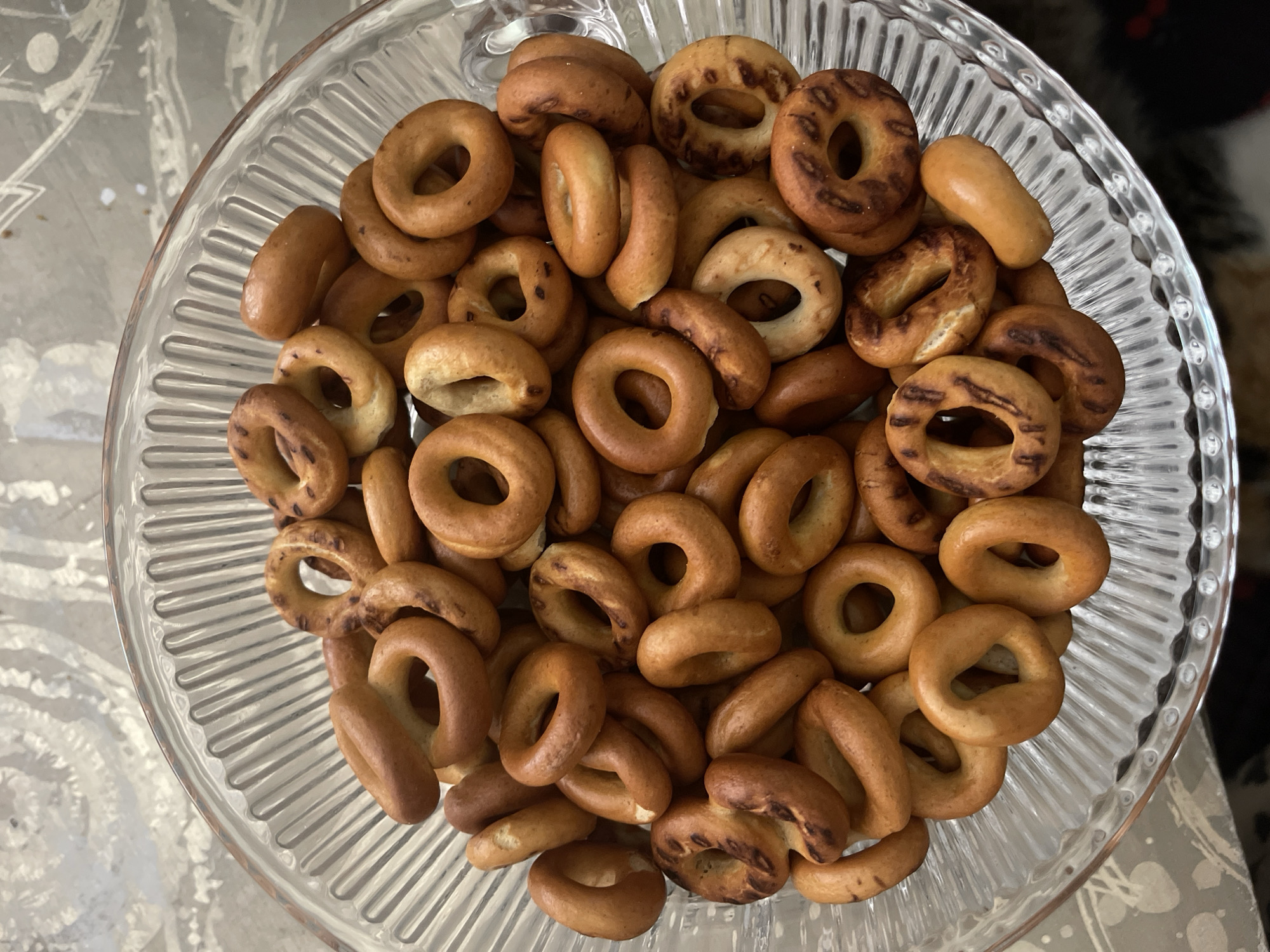 A clear glass bowl filled with small, ring-shaped cookies, commonly known as "baranki" or "sushki" in Russia, is presented from a top-down perspective. The cookies are golden brown in color, with some exhibiting darker, more toasted areas. They vary slightly in size and shape, with most being uniformly circular with a hole in the center, while a few appear more oval or elongated. They are piled generously within the fluted glass bowl, creating a textured and abundant display. The bowl itself rests on a patterned surface, possibly a tablecloth or placemat, with a soft, neutral color scheme. The background is out of focus, suggesting an indoor setting. There is no visible text in the image. The lighting is natural, casting gentle shadows and highlighting the baked goods. The overall impression is one of a simple, home-style treat, perhaps prepared for a casual gathering or as a snack. The image evokes a sense of comfort and tradition associated with Russian cuisine.