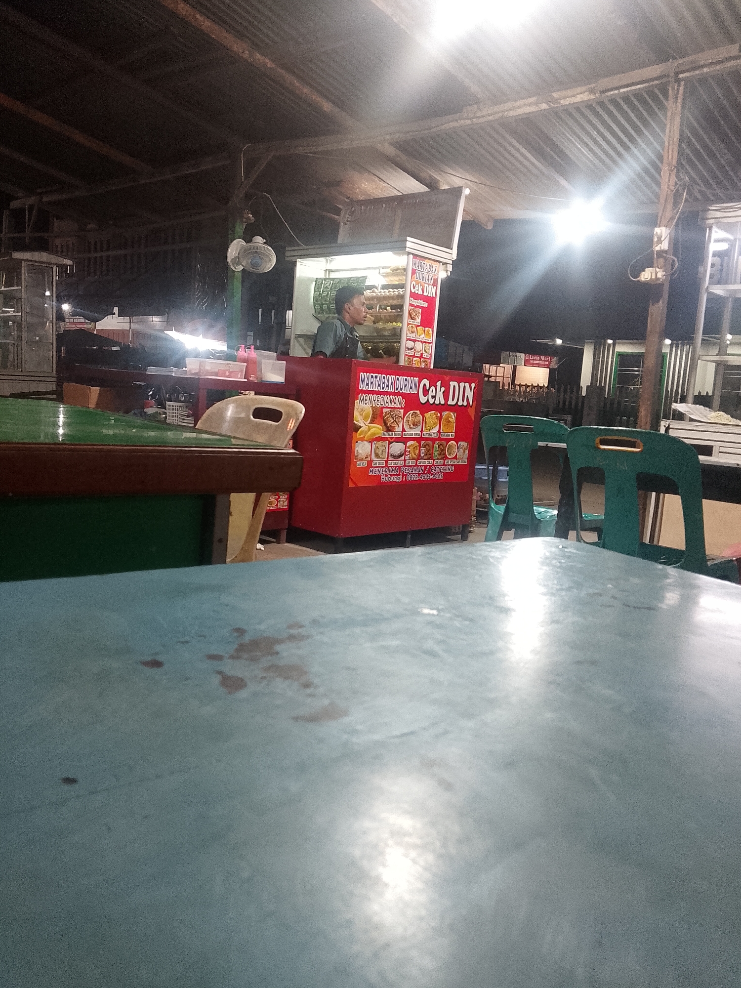 The photo shows a night scene at an outdoor food stall in Lhokseumawe, Indonesia. The stall, marked "MARTABAK DURIAN Cek DIN," offers martabak durian. A person stands inside the stall, which is illuminated by a bright light. In the foreground is a blue table with some stains. A green table and chairs are also visible. The environment is simple with corrugated metal roofs overhead.