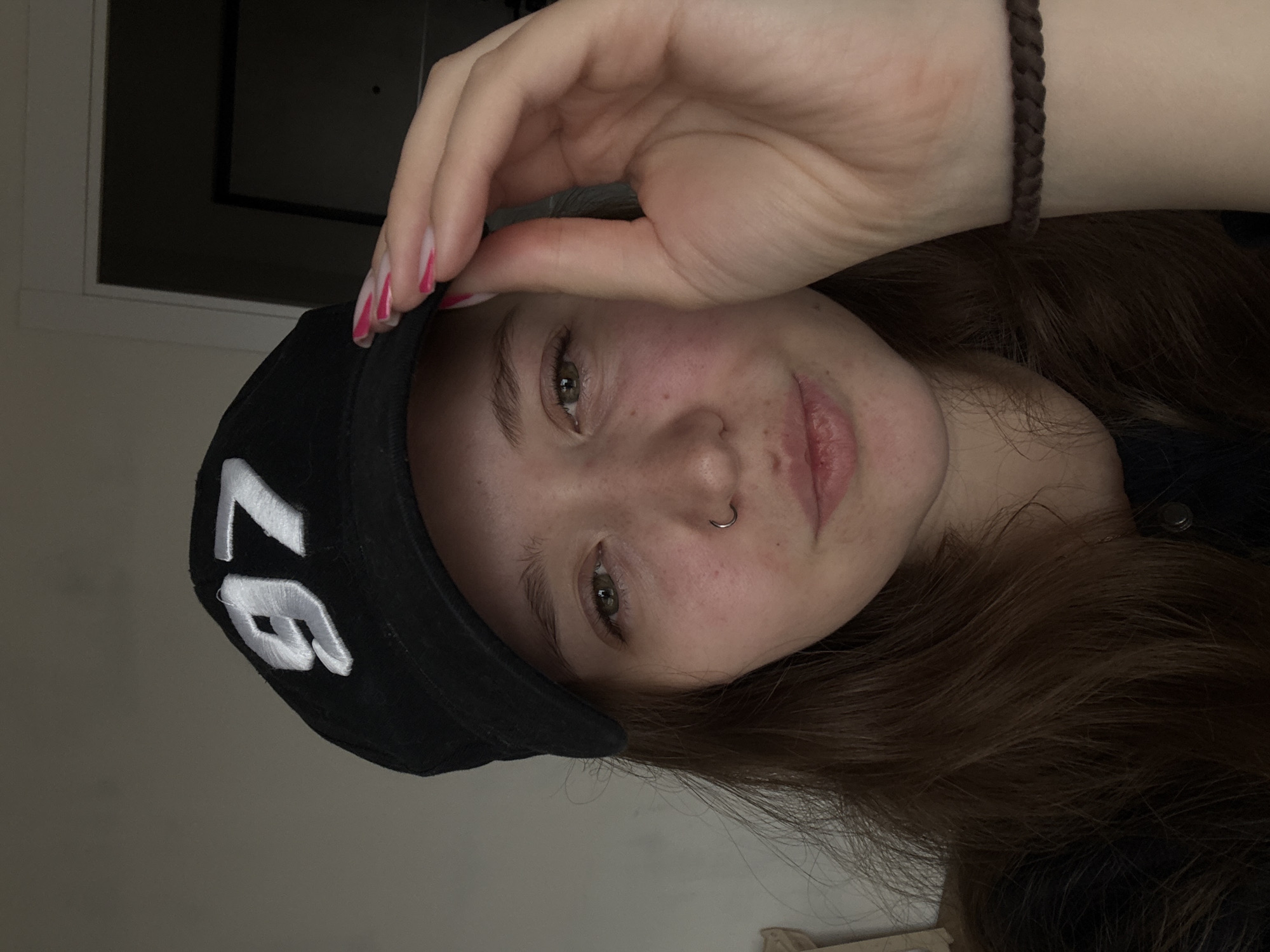 A person with fair skin, light brown hair, and a nose ring is wearing a black cap with white lettering that appears to be "97." The person's left hand is touching the cap. A brown braided bracelet is visible on the person's wrist. The background shows a wall and a partially visible window frame. The image is oriented with the person's head tilted to the right. There is no indication of location in the city of Izhevsk, Russia.