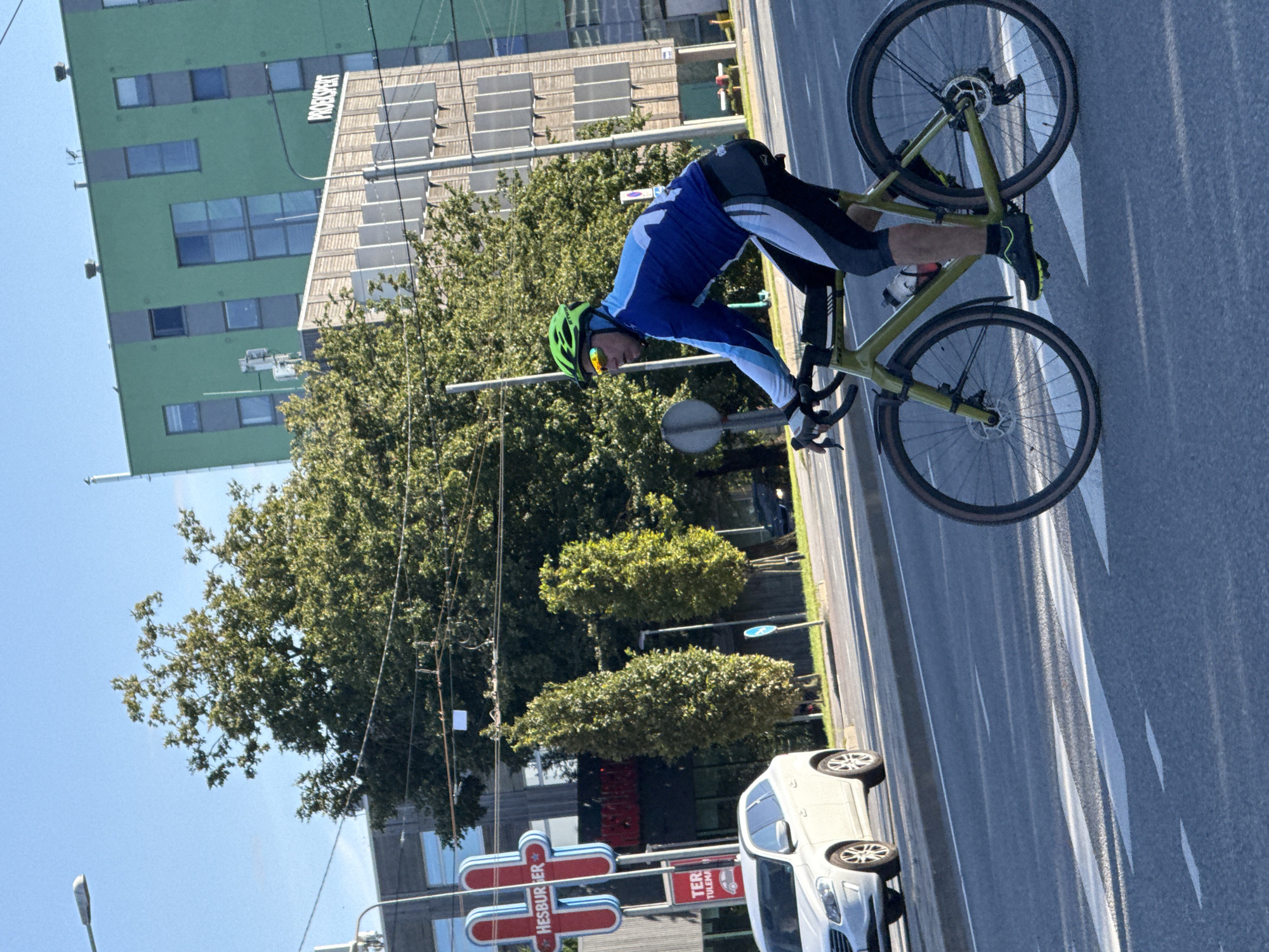A male cyclist wearing a green helmet and a blue and light blue cycling jersey with black and white cycling shorts is riding a green gravel bike across a street in Tallinn, Estonia. The cyclist is facing right and is partially obscuring a white SUV. In the background, a large green building with the word "PROEXPERT" visible is next to a smaller building with a Hesburger sign. Trees and streetlights are also present. The scene appears to be an intersection with pedestrian crosswalk markings on the road. The sky is clear and blue.