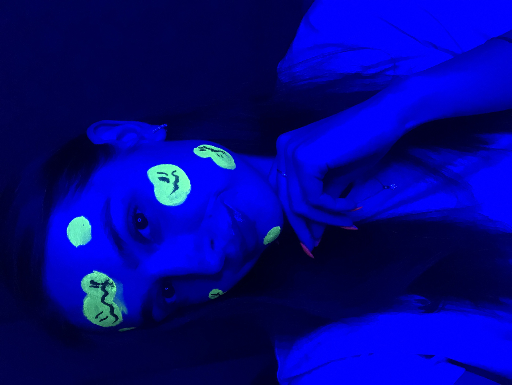 The image shows a young woman with neon green face paint illuminated by blacklight. She has several painted shapes on her face, some resembling smiley faces with wavy mouths and circular eyes. The background is dark, indicating a dimly lit or nighttime environment. The woman is looking towards the camera, and her right hand is raised to her chin, with bright pink fingernails visible. The scene appears to be indoors, given the controlled lighting. There are no discernible elements in the image that would suggest a specific location in Noviy, Russia.