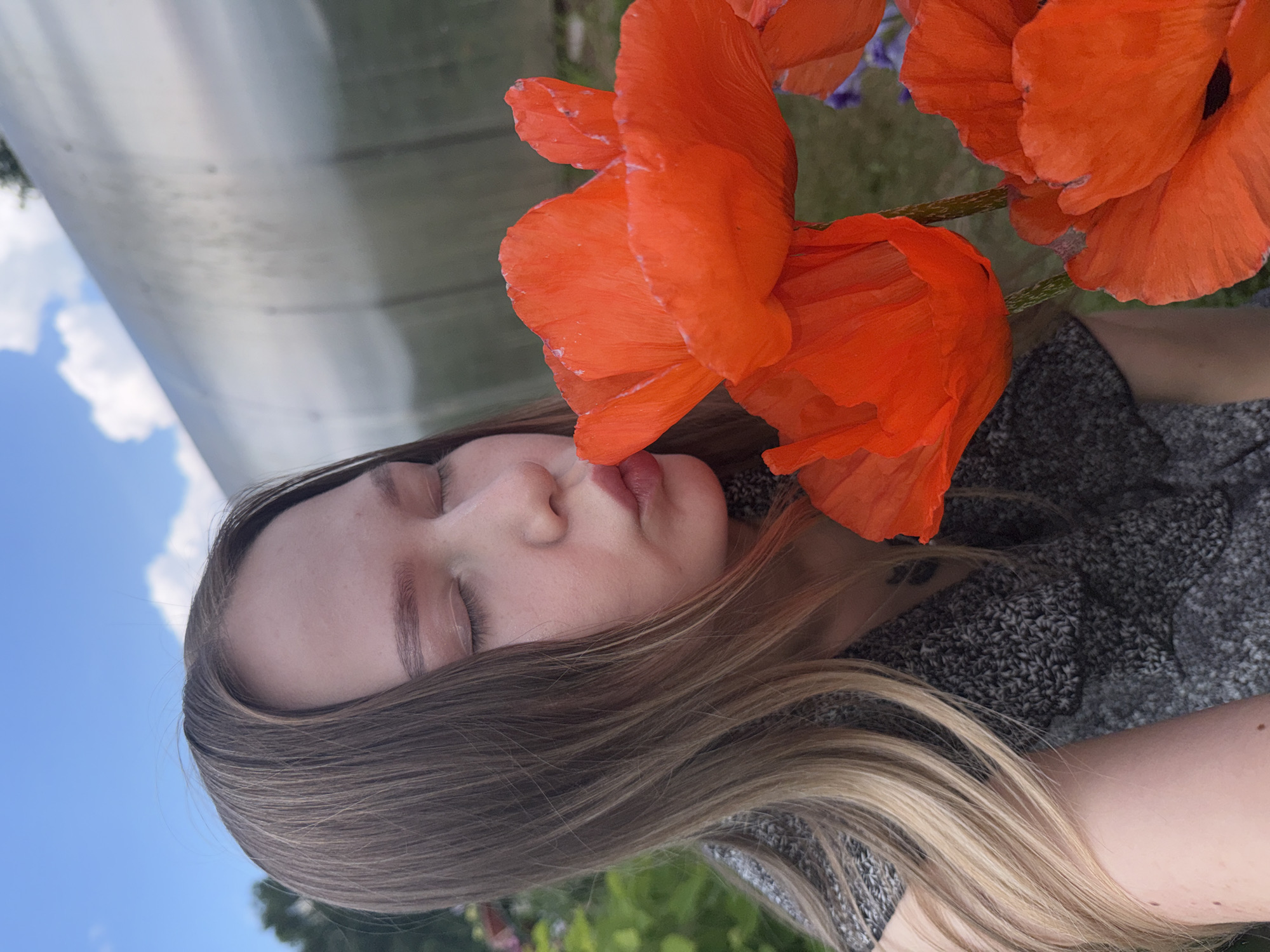In Zhigalki, Russia, a young woman with long, light brown hair and closed eyes is captured in a close-up shot, leaning towards bright orange poppies. Her lips are pursed as if to kiss the vibrant red petals. She is wearing a grey, textured top. The background features a blurry expanse of green foliage and a large, metallic, curved structure, possibly a greenhouse or shed, under a bright blue sky with scattered white clouds. The overall scene is outdoors and appears to be during daylight.