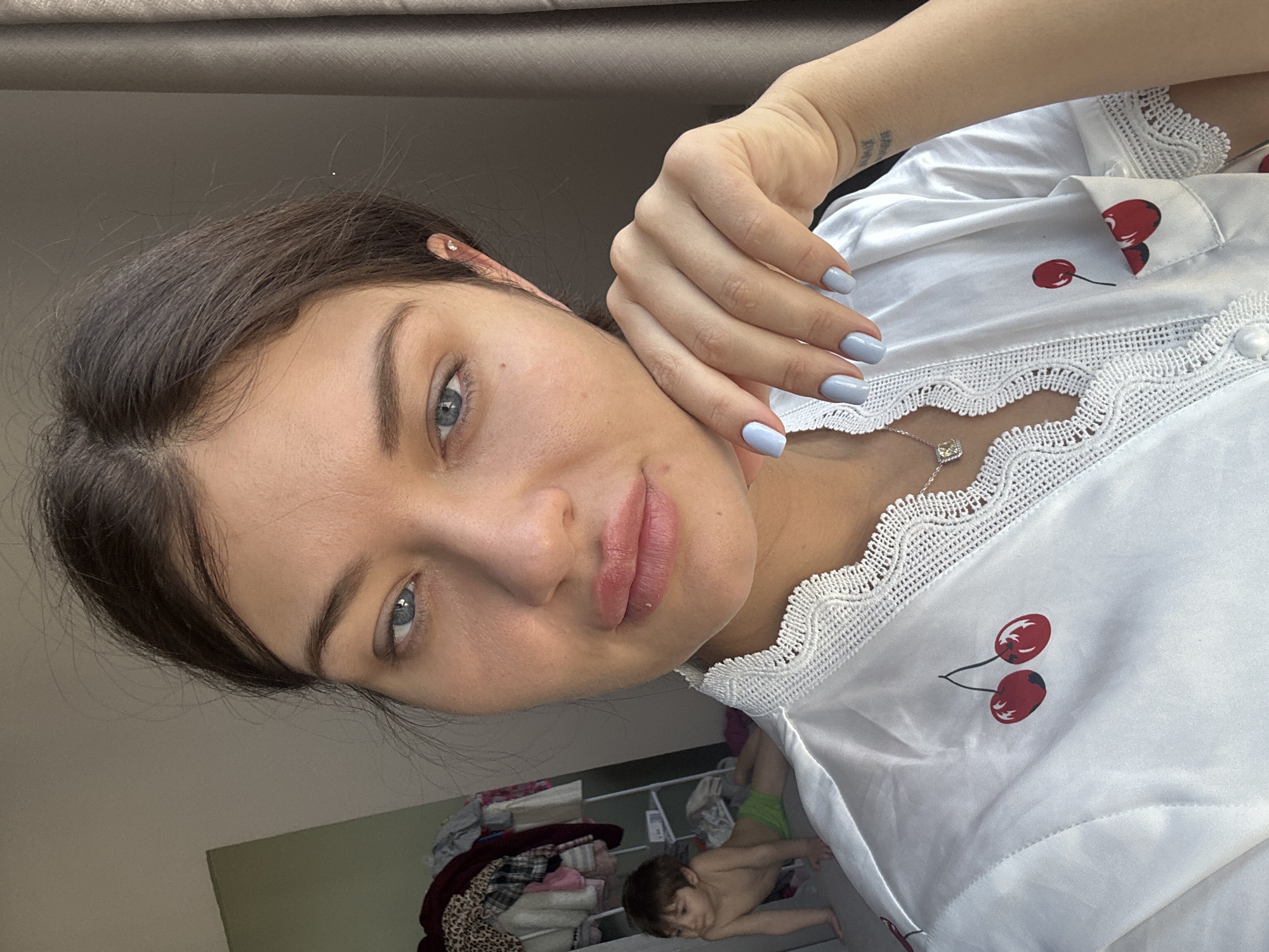 A medium close-up angled portrait depicts a woman with light skin, brown hair, and blue eyes. She has light blue nail polish on her nails and is wearing a white top with red cherry patterns and white lace trim. A silver necklace is visible around her neck. Her left hand is positioned near her face.
In the background, a child is visible lying on the floor, next to a closet filled with clothes. The location is an interior space. There is no information in the image that places the location in Moscow, Russia.